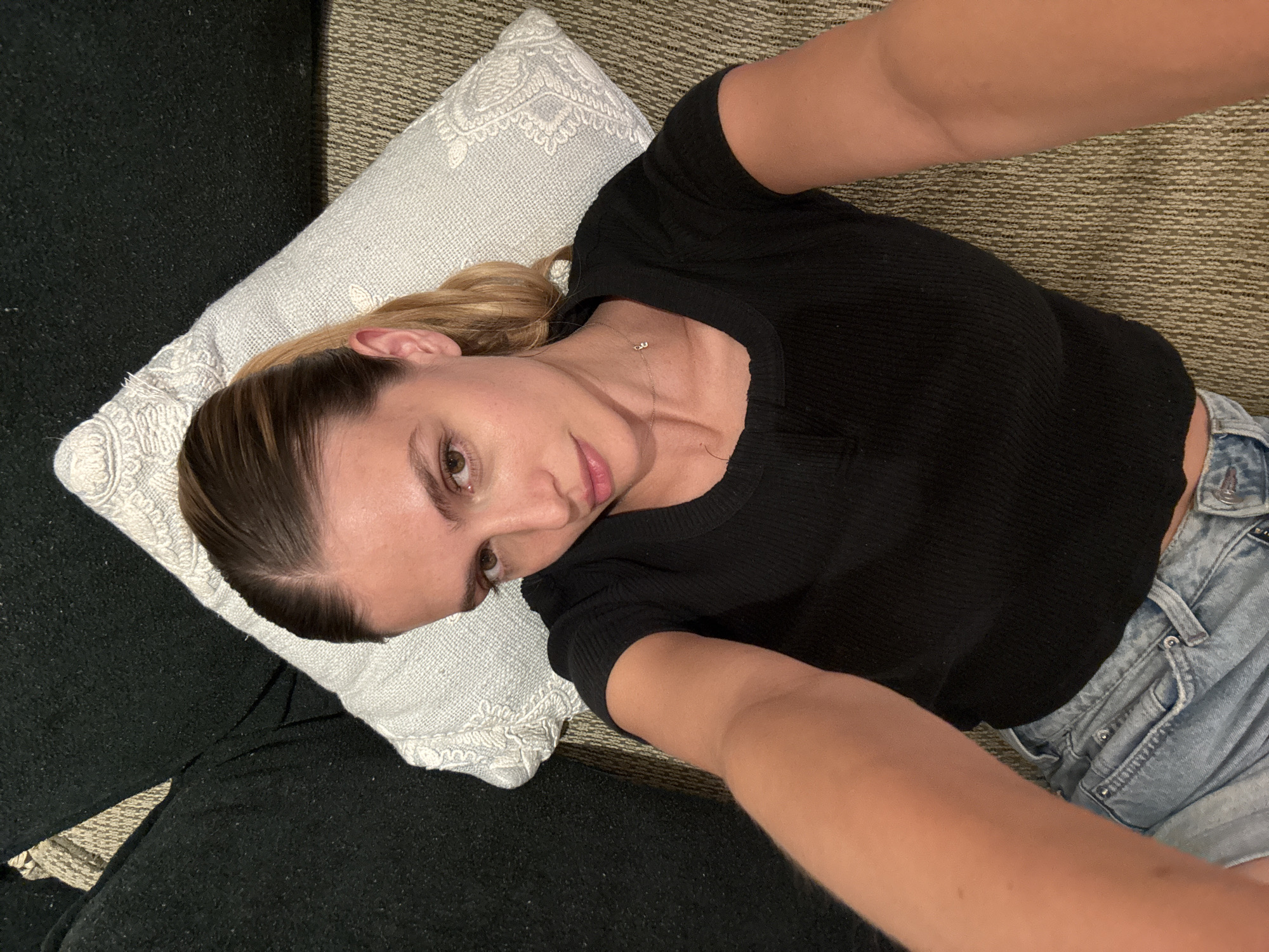 A close-up selfie captures a woman lying down indoors, looking directly at the camera with a neutral and relaxed expression. She has light brown hair pulled back, brown eyes, and light pink lips. She is dressed in a black, ribbed short-sleeved t-shirt and light wash denim shorts featuring silver button details. Around her neck, a delicate gold chain holds a small pendant, which appears to be two small letters, possibly "DU" or "D&J."
Her head rests on a white, rectangular pillow decorated with an intricate, possibly embroidered, pattern. To her left, a dark, likely black, upholstered surface, possibly a couch armrest or cushion, is visible. The background is a textured rug or carpet in shades of brown and beige. The overhead perspective indicates the photo is a selfie. The soft, even lighting suggests an indoor setting, potentially during the evening. The overall atmosphere conveyed is casual and calm.