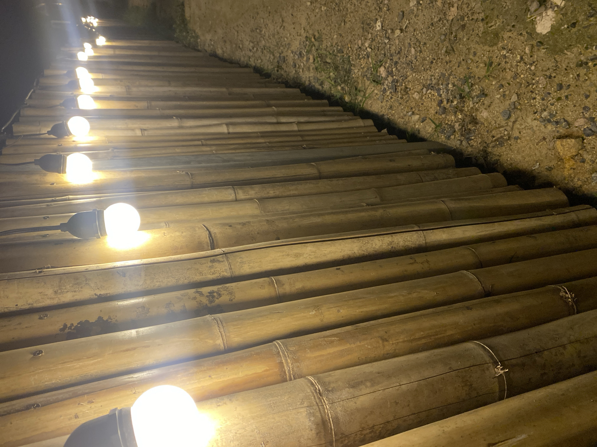 The image depicts a nighttime scene in Baan Don Sai, Thailand. A pathway is constructed of numerous bamboo poles laid side-by-side, creating a walkway. A string of lights illuminates the path, with each bulb emitting a warm, bright glow. The ground on one side of the pathway is dirt and gravel, and the opposite side fades into darkness. No people are visible. The focus is on the illuminated path and the construction materials used.