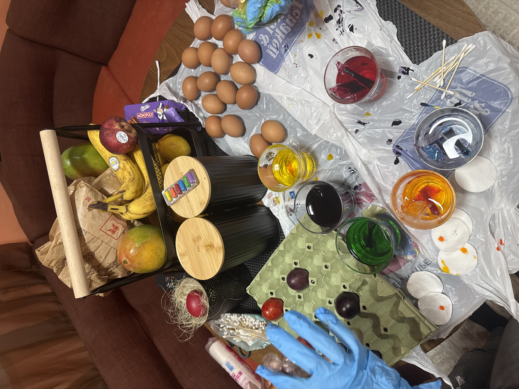 The image shows a still-life composition on a surface, potentially a table, with a mixture of items related to food preparation and Easter egg dyeing.
Objects present:
- A collection of approximately 20 brown eggs.
- A black metal basket containing fruit: bananas, a mango, an apple, and a lemon. A rolling pin is in the basket, as well as a brown bag. There is also a package of Milka Monopoly chocolate in the basket.
- Several cylindrical containers with wooden lids.
- An egg carton holding some dyed eggs in shades of red and purple.
- Several glasses containing colored dyes: red, blue, yellow, black, and green.
- Cotton swabs and cotton pads.
- A blue gloved hand.
- An egg in a decorative nest.
- A white plastic sheet.
Actions/Interactions:
- Someone is likely in the process of dyeing eggs.
Environment:
- The image is taken indoors, possibly in a home. A brown couch is visible in the background.
Location:
- The Kaufland logo is visible. Kaufland is a German hypermarket chain, but does not inherently identify the city of Sofia, Bulgaria. The specific city in which the image was taken is undetermined without further information.