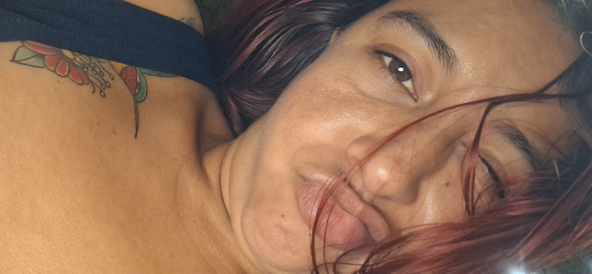The image is an intimate, close-up portrait of a person, likely female, illuminated by soft, natural light. The subject is seen from a slightly elevated perspective, lying down with their head tilted. One brown eye is visible, directly engaging the viewer with a relaxed, perhaps contemplative or sleepy expression, while the other eye is partially obscured by reddish-brown hair that cascades across the face. The person's lips are slightly parted, and their skin has a tan complexion, showing natural textures and some fine lines around the eye.
A blue strap, possibly from a top or garment, is visible on the person's right shoulder and upper back. Peeking from beneath this strap is a colorful tattoo. The tattoo features floral elements with prominent red petals, a yellow center, and green leaves, along with what appears to be part of a small, stylized bird, possibly a hummingbird. The overall atmosphere of the image is serene and personal. The time of day appears to be daytime, given the natural light. There are no specific location cues visible within the frame, but the provided context for the image is Vallecito, Venezuela. No text is visible in the media.