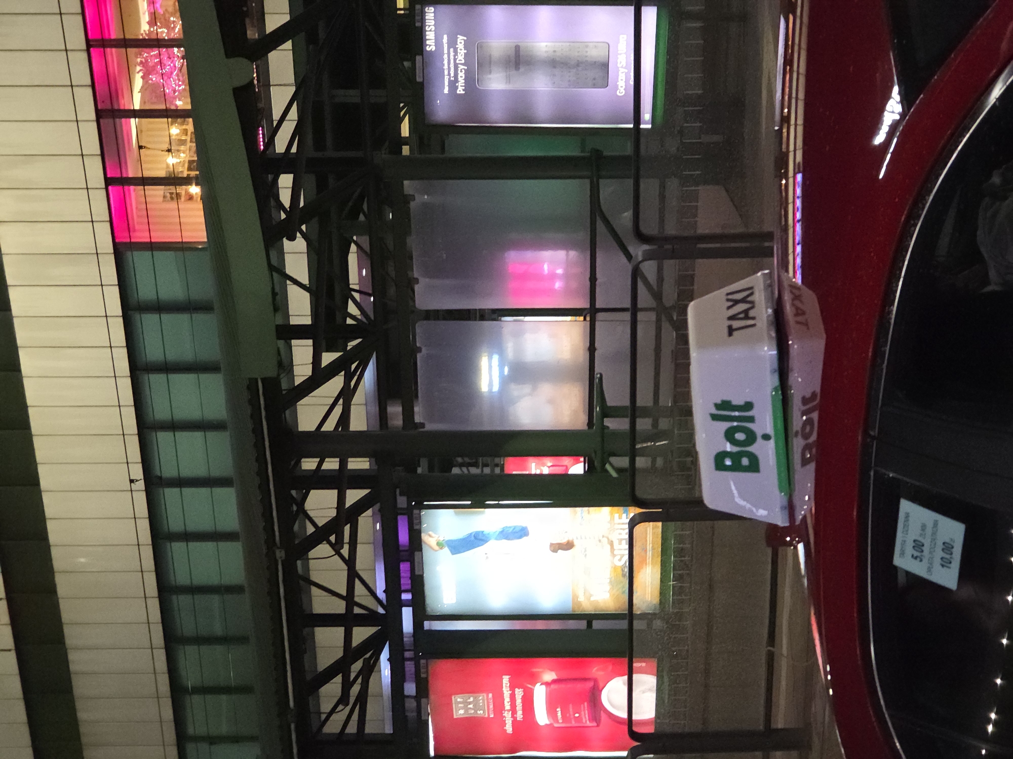 The image depicts a night scene in Warsaw, Poland, featuring a red "Bolt" taxi parked near a building with multiple illuminated advertising billboards. The billboards showcase various products and brands, including Samsung's "Privacy Display" for the Galaxy S23 Ultra, and products from "Ritual" and "Sierie." The building behind the taxi has a structure of metal beams and columns, with lights and decorations visible within. The taxi has a roof-mounted "Bolt" sign and a sticker on the window displaying fare information.