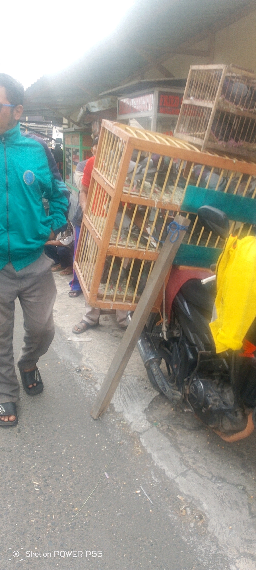 A man in a teal jacket and gray pants stands on a sidewalk next to a wooden cage filled with pigeons. The cage is balanced on the back of a motorcycle. Another man in a red shirt is partially visible behind the cage. In the background, there is a food stall with a red sign that says "FRIED CHICKEN". The ground is paved with asphalt and some debris. The overall scene suggests a street market or a roadside vendor.