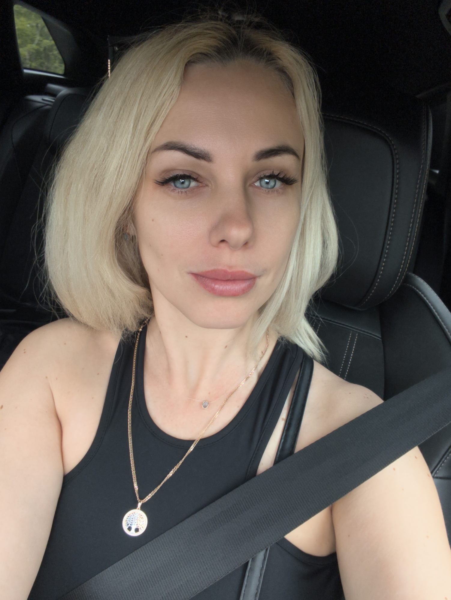 A light-skinned woman with blonde hair and blue eyes is sitting in the driver's seat of a car. She is wearing a black tank top and a seatbelt. She also has two necklaces, one with a tree-of-life pendant. The location cannot be identified as Novorossiysk, Russia, from the image alone.