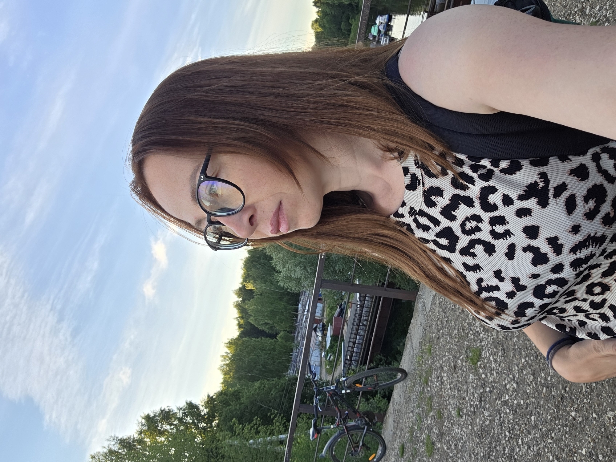 A woman with long brown hair and glasses is pictured from the chest up, looking to her right, outdoors. She is wearing a sleeveless top with a leopard print pattern and a black backpack. In the background, a bicycle is leaning against a railing. Beyond the railing is a body of water with several boats, and a densely wooded area. The sky is blue with scattered white clouds. The scene appears to be a scenic spot near a body of water, possibly a park or recreational area. This appears to be the embankment of the Kama River in the city of Chaikovsky, Russia.