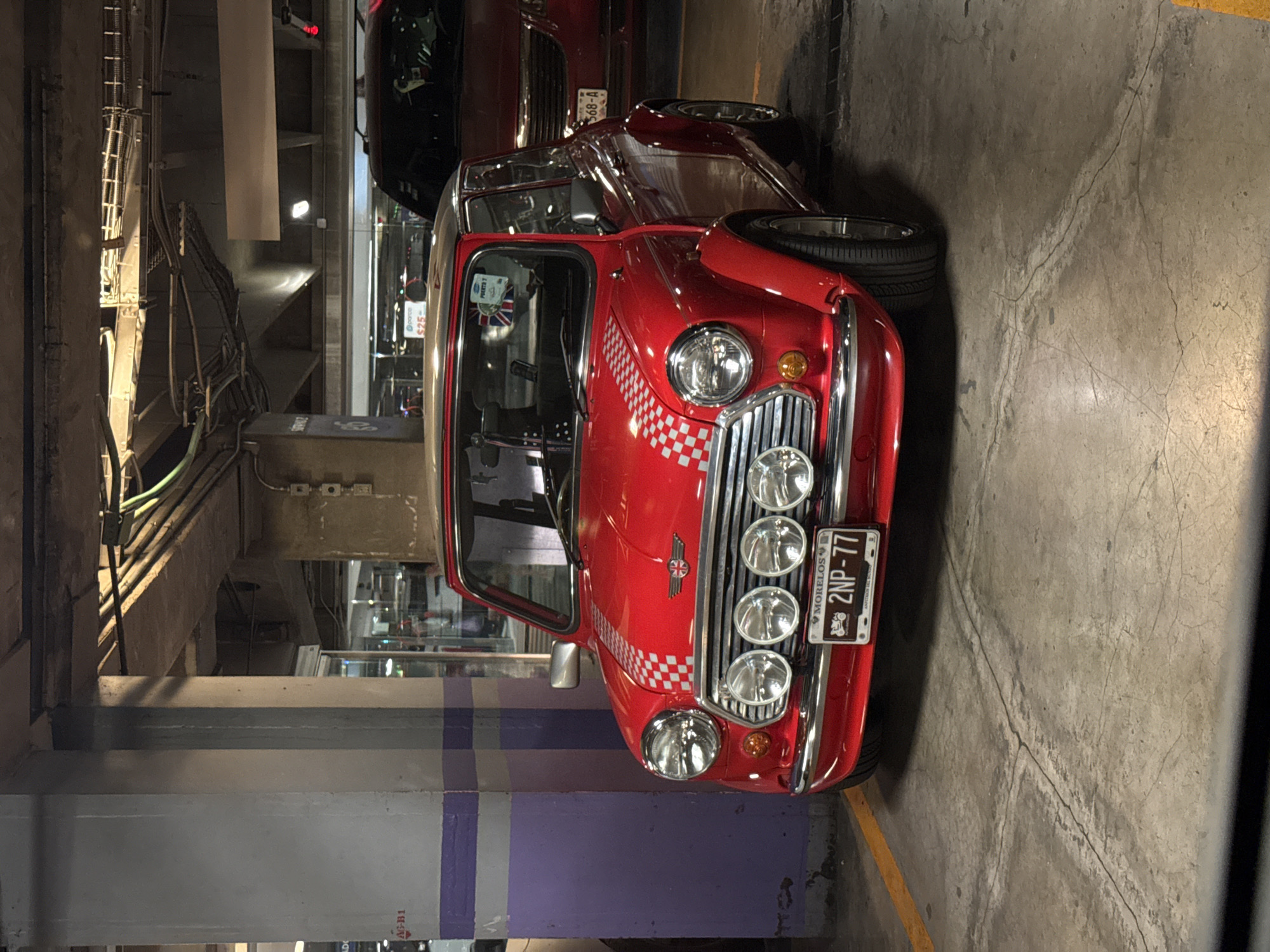The image shows a red classic Mini Cooper parked in a parking garage in Mexico City. The car has a distinctive checkerboard pattern on the hood and a license plate from Morelos, Mexico reading "2NP-77". The garage is concrete, with visible support beams painted with purple and gray stripes. Other cars are visible in the background. The setting is urban, suggesting an indoor parking structure within Mexico City.