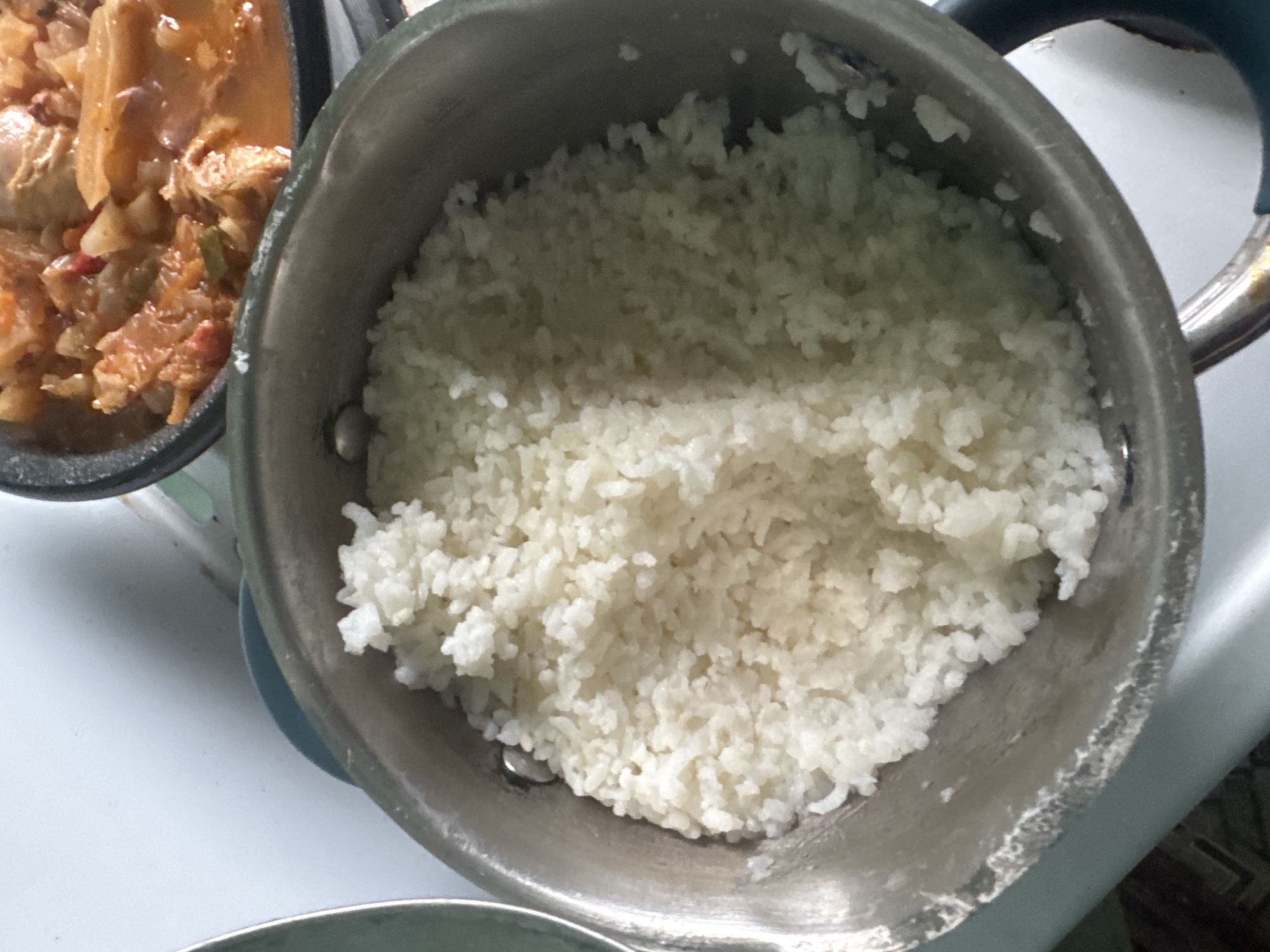 A close-up, overhead shot captures a pot filled with fluffy white rice. The pot is silver-toned and shows signs of use. To the right, a portion of another pot is visible, containing a hearty stew or curry with visible pieces of meat and vegetables in a reddish-orange sauce. The background appears to be a kitchen counter. The image does not contain information to determine the location within the city of Krasnoe, Russia.