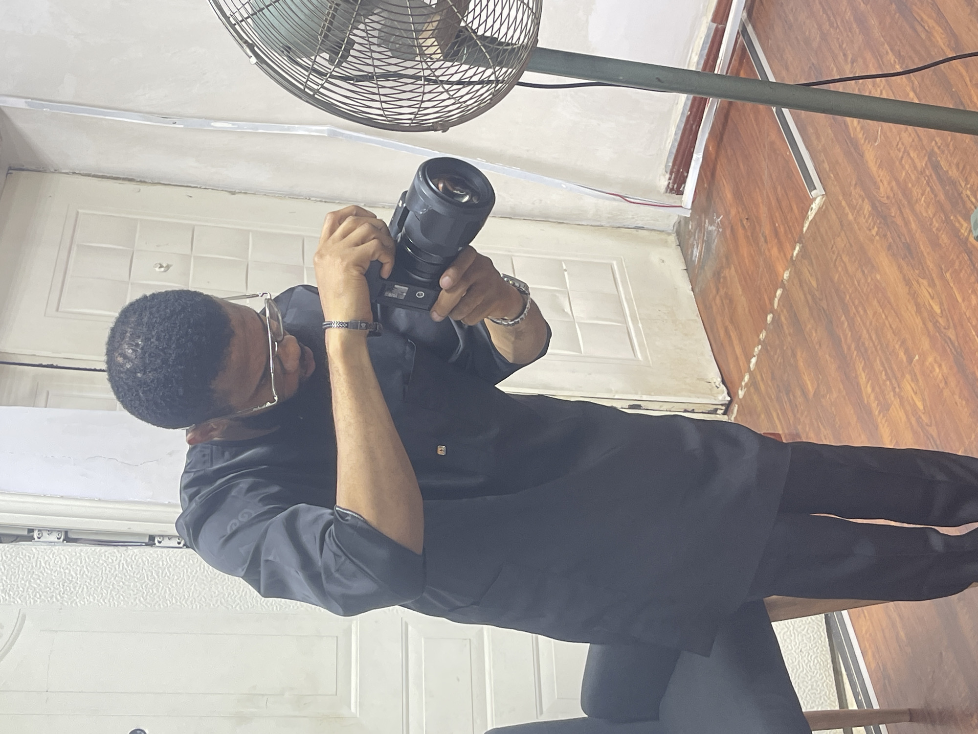 A man is holding a camera indoors, likely in a residential or office space, based on the presence of a door, flooring, and a fan. The man is wearing glasses and dark clothing. The room appears to be in a building in the city of Abuja, Nigeria.