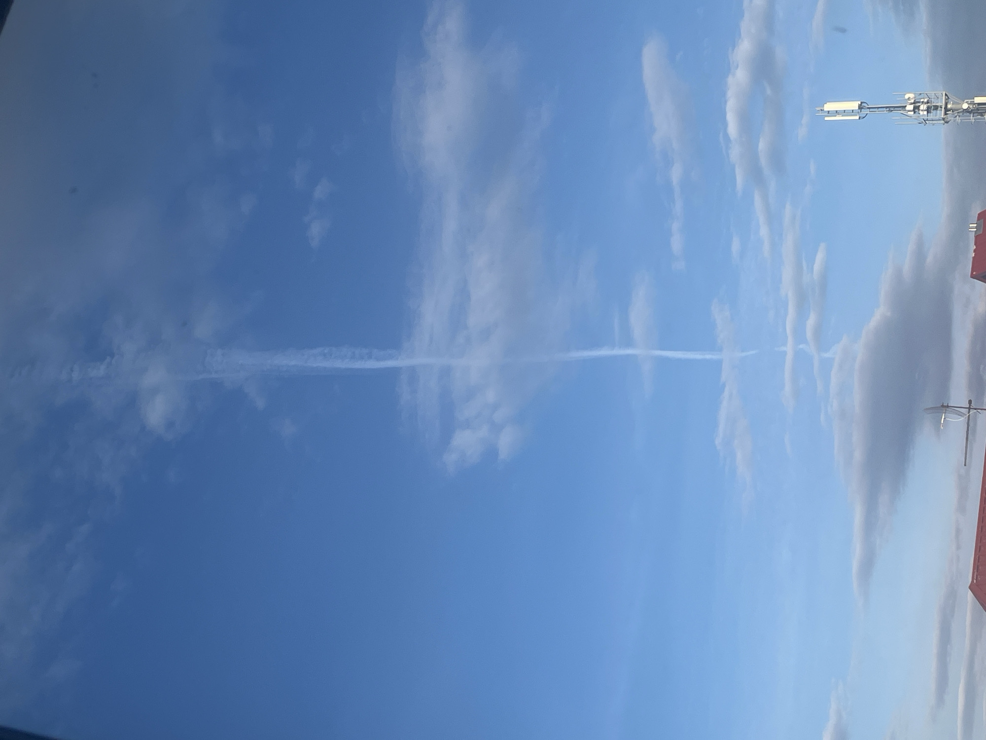 A wide-angle, daytime photograph captures a bright blue sky with scattered clouds and a prominent contrail. The image appears to be taken looking upwards, likely from a ground-level or rooftop perspective in Ulyanovsk, Russia.
A long, thin, white contrail, presumably from a passing jet aircraft, stretches horizontally across the middle of the frame, dividing the expanse of the sky. The contrail is distinct but shows signs of diffusion at its edges, gradually blending into the surrounding atmosphere.
The sky is a vibrant azure blue, indicative of clear weather conditions. White, wispy cirrus clouds and some larger, light gray cumulus clouds are dispersed across the sky, particularly on the right side and near the bottom of the frame (when viewed in standard landscape orientation).
On the right side of the image, two man-made structures are partially visible, suggesting an urban or suburban setting. At the upper right, part of a modern cell tower or communications mast is seen, featuring multiple white rectangular antenna panels and metallic support beams. Below this, the reddish-brown roof of a building is visible, with a small, white satellite dish antenna mounted on a pole, and another smaller, rod-like antenna structure.
No people or discernible text are present in the image. The bright illumination suggests it is mid-day or early afternoon.