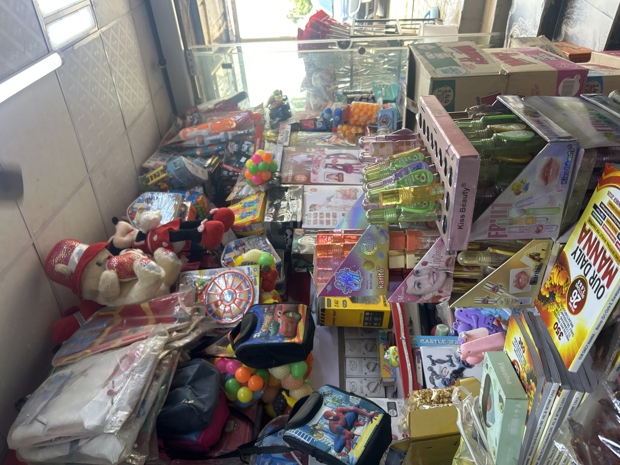 This is a full-shot, eye-level, medium-width image taken inside a small shop in Dutsen Alhaji, Nigeria, likely a convenience store or a small market stall. The scene is densely packed with a variety of merchandise, indicating a bustling retail environment.
In the foreground, on the right side, there is a stack of cardboard boxes. On the left, a pile of books with yellow covers prominently displays the title "OUR DAILY MANNA" and "350 Preaching Prayers". A small, green tissue box with an avocado illustration sits atop these books, next to a pink and purple toy figure. Further left, a transparent plastic bag contains dark, dried meat-like items.
Dominating the center of the image is a display rack filled with numerous small bottles of lip gloss or oil, some with green liquid and others with purple. The brand "Kiss Beauty" is visible on the packaging. Behind these, more stacked boxes and various toys are visible, including a yellow toy car and bags featuring cartoon characters like Spider-Man and characters from the movie "Cars".
The upper section of the shop is filled with more merchandise hanging from the ceiling or stacked on shelves. These include large plush toys like a teddy bear wearing a sequined red hat and Mickey Mouse, as well as various packaged goods, balloons, and kites. The ceiling is made of white, patterned tiles, and two fluorescent lights are visible.
The background shows the entrance to the shop with a glass door and a glimpse of the outside environment, suggesting an urban or semi-urban setting. A silver car is partially visible through the glass. The overall impression is one of an organized yet crowded retail space selling a diverse range of items, from personal care products to toys and religious literature. There are no people visibly present in the scene.