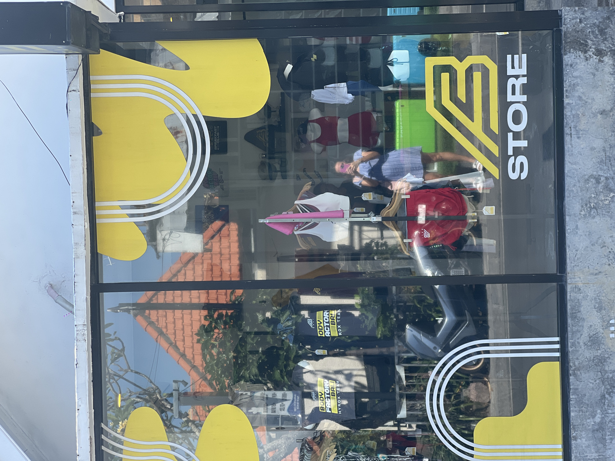 A storefront in North Kuta, Indonesia, displays a glass window with black framing. The window is partially covered with a yellow design and white parallel lines. On the right side of the window is a large yellow and white logo with the text "STORE" next to it. The reflection shows a red-tiled roof of a building across the street, while within the store, various items of clothing are on display. Inside the store, a person wearing a light blue dress and a pink face mask is visible. Part of the logo includes a stylized "B." The storefront is also partially obscured by branches and leaves.