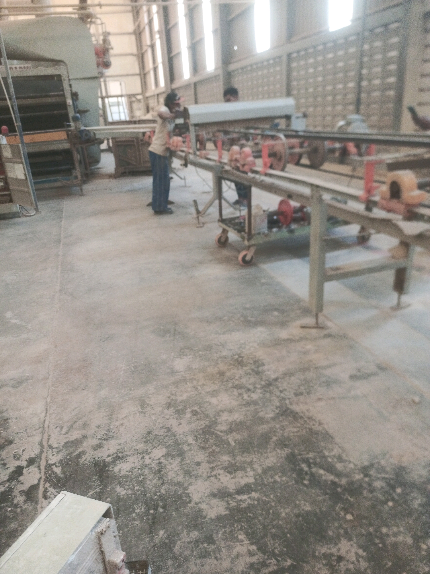An indoor industrial setting in Lebaniwaras, Indonesia, shows two individuals working near a conveyor belt system. The floor is concrete and dusty. To the left, a large, grey industrial machine with multiple shelves is visible. In the center, a person wearing a tan shirt and blue pants stands next to a conveyor belt, appearing to be engaged in work. Further to the right, a second person is partially visible behind the conveyor belt. The conveyor belt system consists of metal rails and various components, including red and orange parts, suggesting an active manufacturing or processing area. The environment is lit by natural light coming through windows or openings in the building's walls, which are constructed with a brick-like pattern.