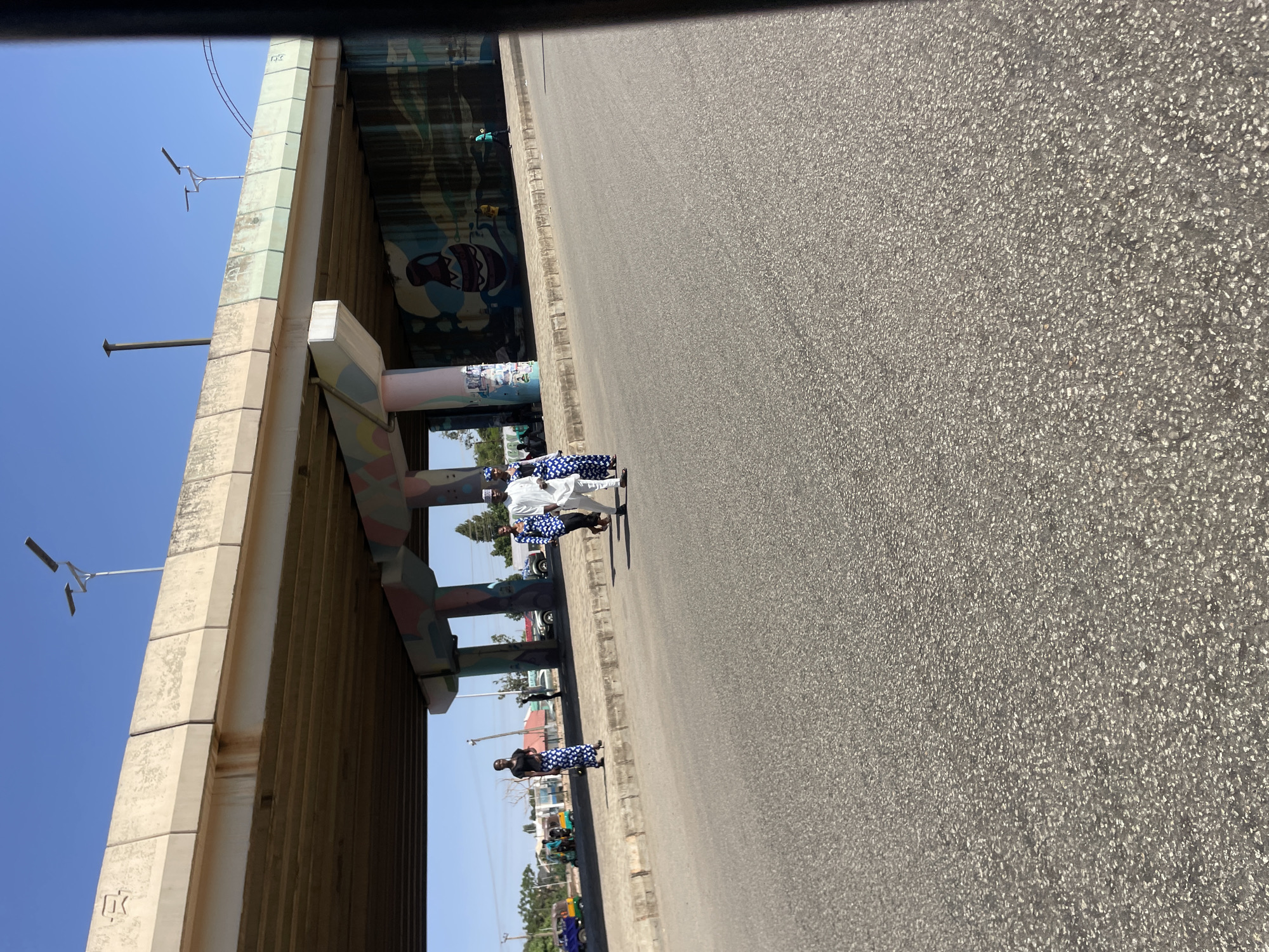 A partially obstructed view of a road running parallel to a concrete overpass in Jos, Nigeria, is seen in the image. The right side of the image is largely comprised of a gray asphalt road. A concrete overpass structure occupies the left side, featuring graffiti and support columns in light colors. Several individuals are standing along the road's edge, adjacent to the overpass; most are wearing white and blue patterned clothing. The sky is clear and blue. In the distance, trees and other buildings are visible. The photograph is angled.