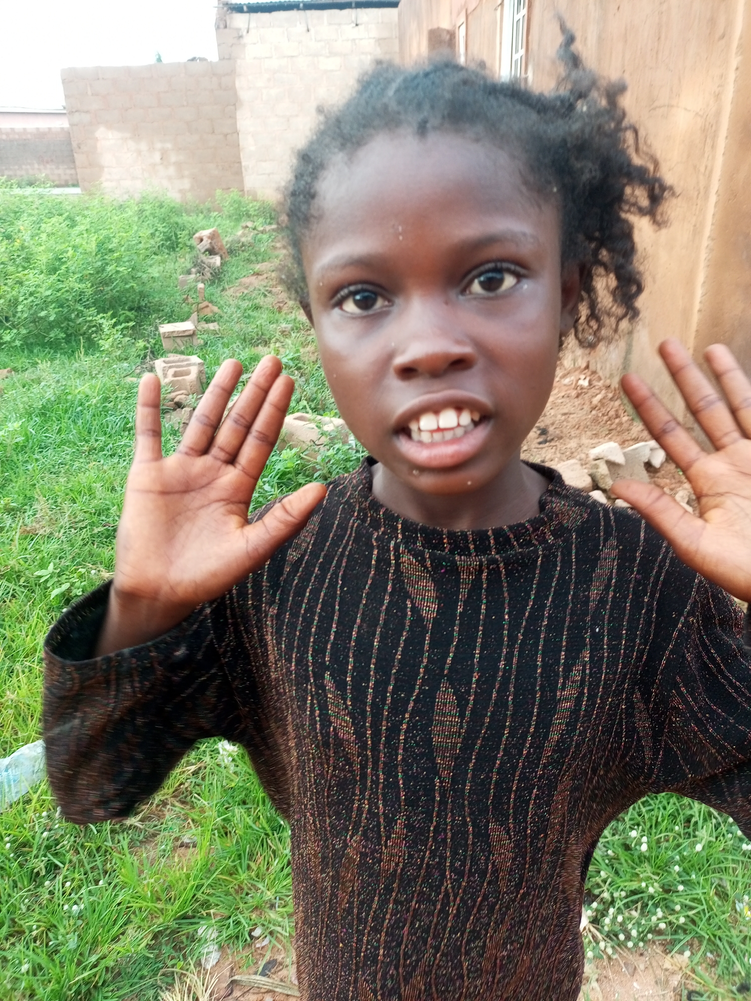 A young girl is pictured outdoors in what appears to be a residential area in Tempure, Nigeria. She is facing the camera, her hands raised and open in front of her. Her hair is dark and curly, and she is wearing a black sweater with thin, vertical metallic stripes. The background consists of green grass, dirt, and a light-colored wall of a building. There are also some loose bricks visible in the grass. The lighting suggests it is daytime.
