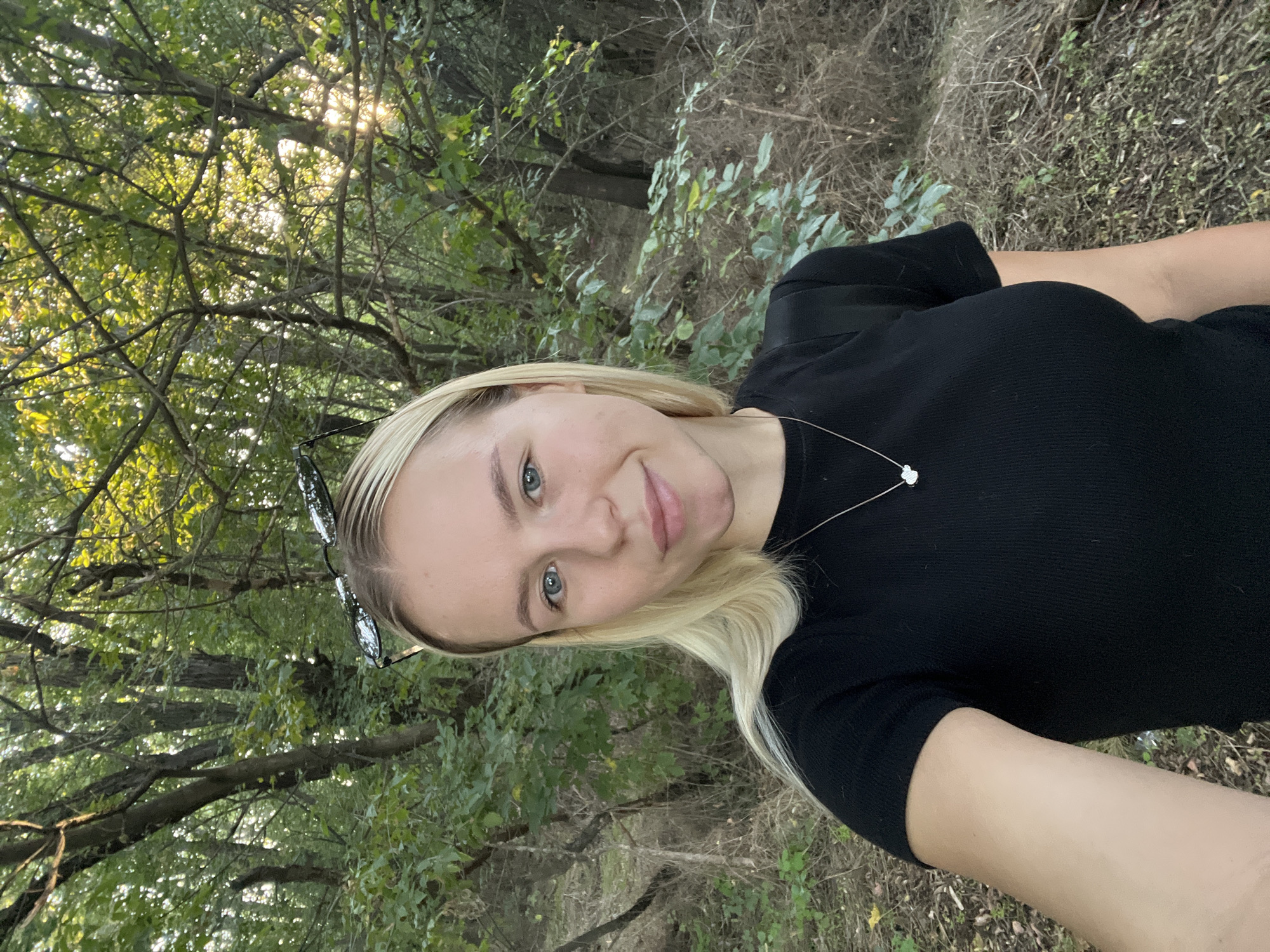 A young woman with blonde hair and blue eyes is taking a selfie in a wooded area. She is wearing a black t-shirt and a necklace with a small clover pendant. Sunglasses are perched on top of her head. The background is filled with trees and foliage, with sunlight filtering through the leaves. The ground is covered with fallen leaves and dry grass.