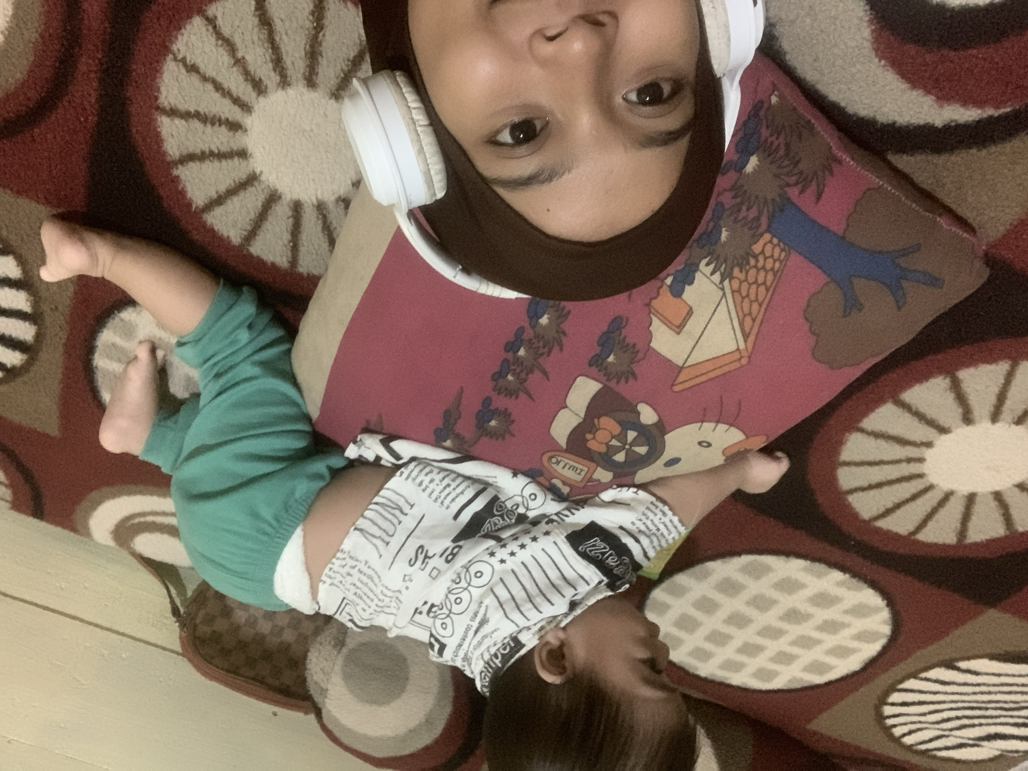 The image captures a selfie taken from a low angle, featuring a baby and a woman in the foreground, with a patterned carpet and a wall in the background. The baby, positioned in the upper half of the frame, is lying on its stomach, wearing a white shirt with black text and patterns, and green pants. The baby's face is not fully visible, but its body is turned to the side. In the lower part of the frame, a woman is looking directly at the camera. She is wearing a brown hijab and white headphones. Her face is the primary focus of the foreground. The environment appears to be indoors, with a rug featuring circular and diamond patterns. The background includes a section of a wall and a bag with a checkered pattern. The image's composition suggests a casual, domestic setting. There is no information in the image to indicate its location in Sungai Meriam, Indonesia.