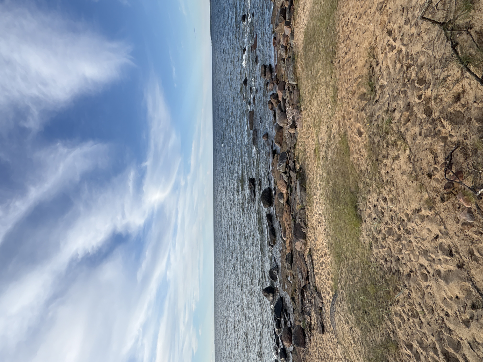 The image shows a serene natural scene of the Baltic Sea coast in Paldiski, Estonia. The foreground is dominated by a sandy beach with scattered dry grass and some fallen branches. The sand is textured with footprints and small stones. Beyond the beach, a rocky shoreline extends into the water, with numerous dark-colored boulders of various sizes partially submerged or resting on the wet sand. The sea itself is a deep blue, with gentle waves creating white foam as they meet the rocks. In the distance, the horizon is a clear line where the sea meets the sky. The sky is a vibrant blue, adorned with wispy white clouds that stretch across the frame in streaks, suggesting a dynamic yet calm weather pattern. The overall impression is one of a peaceful, natural coastal landscape.