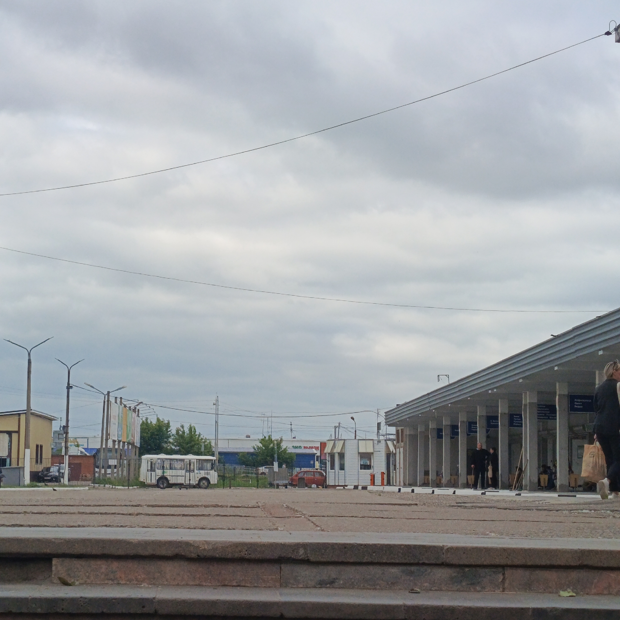 The image depicts a cloudy day at what appears to be a bus station or transportation hub in Chaikovsky, Russia. In the foreground, there are concrete steps leading up to a paved area with rows of what could be parking spaces or waiting areas. To the left, a yellow and brown building with a few windows is visible. Further back, a white bus with blue accents is parked. Across from the bus, a red car is parked near a small white building with an awning.
Dominating the middle ground and extending to the right is a covered walkway supported by numerous pillars, characteristic of a station's waiting area. There are signs with text hanging from the pillars. Several people are present within this covered area, some standing and others seated. One man in a dark suit stands with a cane. A woman in a dark jacket and white shoes walks towards the right edge of the frame, carrying a large paper bag. The sky is overcast with diffuse clouds. Streetlights are visible to the left, and power lines crisscross the sky.