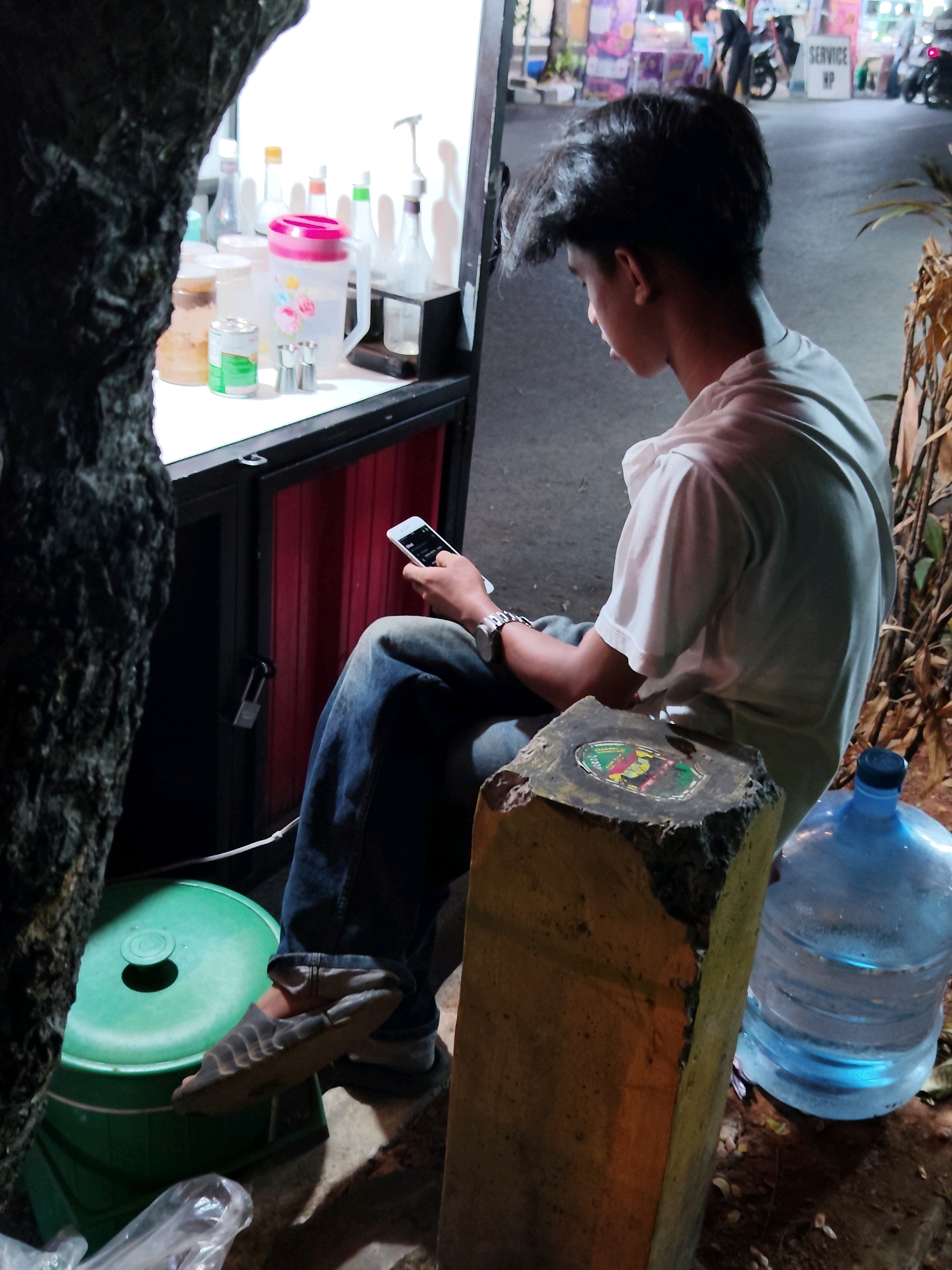 The image depicts a young person sitting on the ground in front of a stall or shop in Purwakarta, Indonesia. The person, with dark, messy hair, is wearing a light-colored t-shirt and blue jeans, and is looking down at a smartphone held in their right hand. A watch is visible on their left wrist.
To the left of the person, partially obscured by a dark, textured object that appears to be a tree trunk, is a green plastic tub with a dark circular indentation on its lid. In front of this tub, a clear plastic bag is visible on the ground.
Behind the person is the stall or shop area, with a white countertop displaying various bottles and containers. These include a large pink-lidded pitcher with a floral design, a tin can, and several smaller bottles. A dark-colored cabinet with red wooden doors is visible below the countertop, secured by a padlock.
To the right of the person, a large blue water jug is prominently displayed on the ground. In front of the water jug and the person is a rough, weathered concrete block or marker. The background shows a street scene with lights and other stalls or shops, suggesting an urban commercial area. The overall lighting suggests it is evening or night.