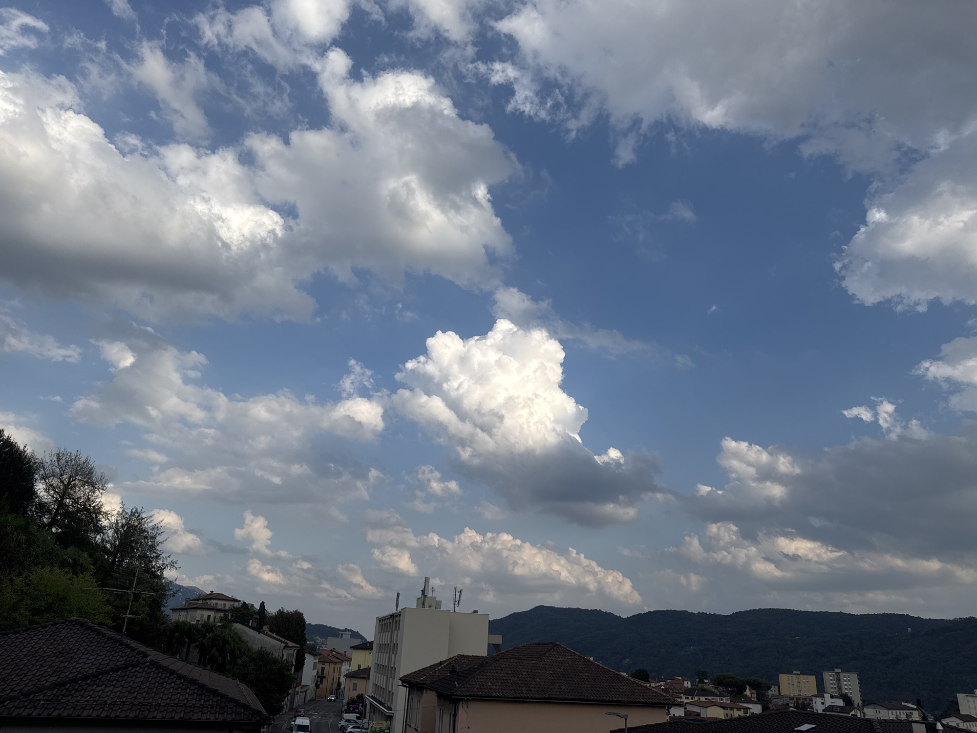 The image depicts a suburban landscape under a dynamic sky filled with a mix of fluffy white cumulus clouds and darker, more ominous formations against a vibrant blue backdrop. The viewpoint is elevated, looking down over a residential area.
In the foreground, tiled rooftops of houses dominate the lower portion of the frame. Beyond these, a street is visible with several cars, indicating it is an active urban or suburban area. Buildings of various sizes and architectural styles are scattered throughout the mid-ground, including what appears to be a multi-story office building with antennae on its roof. The buildings suggest a developed settlement.
The background features rolling, tree-covered hills that form a natural boundary to the settlement. The overall impression is of a town or city nestled amongst verdant hills, under a partially cloudy sky. The lighting suggests it is daytime, with sunlight illuminating the clouds and casting some shadows. The specific location is identified as Balerna, Switzerland.