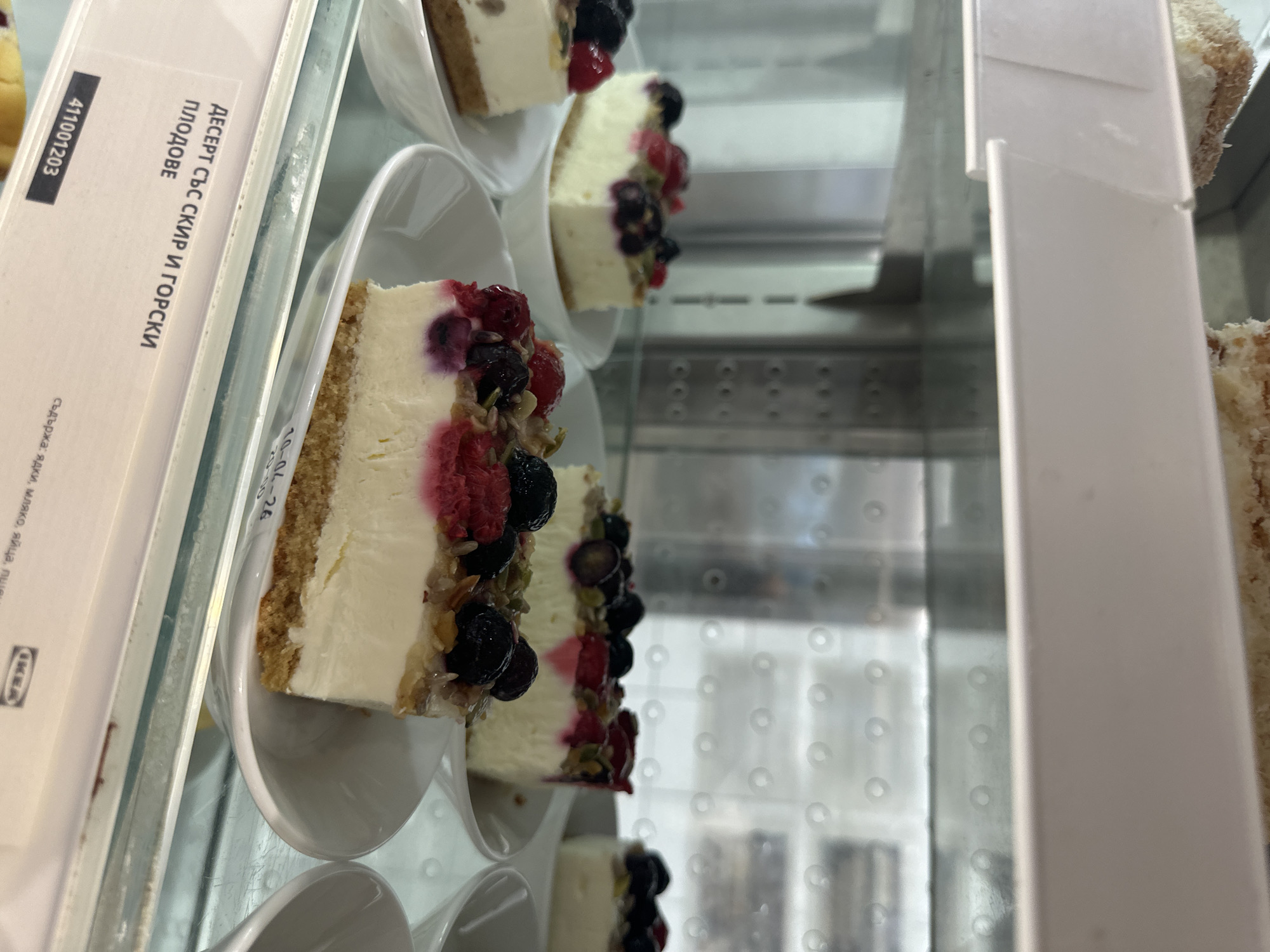 The image shows a close-up view of a refrigerated display case filled with rows of individual portions of a dessert, likely cheesecake, topped with mixed berries. The desserts are arranged on small, white, spoon-shaped dishes. The label visible in the foreground identifies the dessert in Bulgarian as "ДЕСЕРТ СЪС СКИР И ГОРСКИ ПЛОДОВЕ", which translates to "Dessert with skyr and forest fruits". It includes the number "411001203" and the date "10-04-26". The IKEA logo is also visible, implying this dessert is being sold at an IKEA store. Given the Bulgarian language on the label, the location is most likely Sofia, Bulgaria, where IKEA has a presence. The display case itself appears to be made of stainless steel with a glass or acrylic front. The interior has a grid-like pattern, possibly part of the refrigeration system.