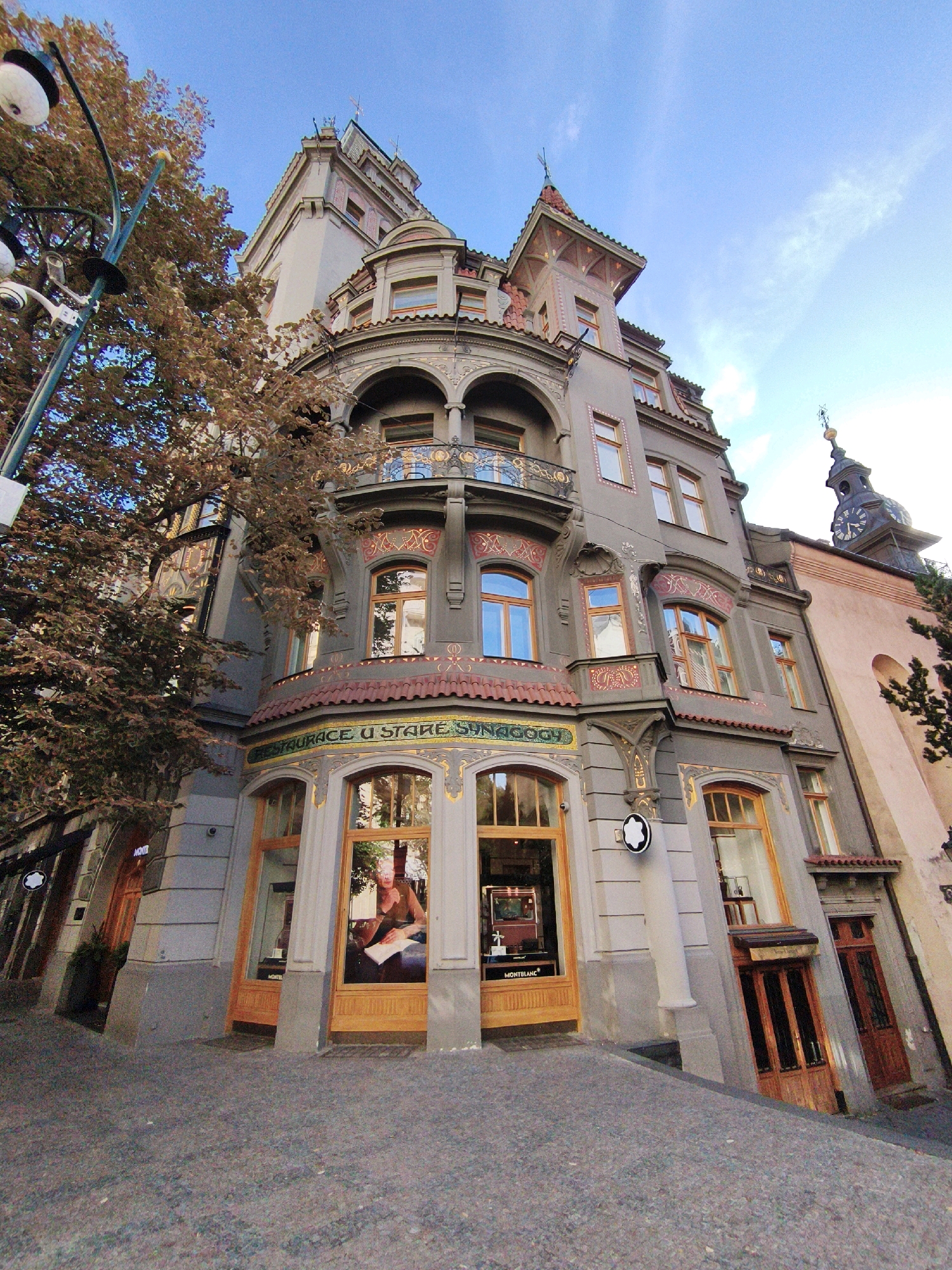 The image shows a low-angle, wide-shot of a building in Prague, Czechia, under a blue sky with scattered clouds. The building is an ornate, multi-story structure with a mix of architectural styles, featuring a rounded facade with arched windows, decorative balconies, and a prominent clock tower. The building's exterior is painted in a neutral gray, with accents of red, gold, and terracotta.
At street level, the building has large display windows, one of which shows a person sitting at a table with a piece of paper. The signage above the windows reads "RESTAURACE U STARE SYNOGOGH." Another window displays a Montblanc product. A large tree with brown foliage partially obscures the left side of the building. The ground in front of the building is paved with cobblestones. A street lamp with two lights and security cameras is visible on the left. The overall scene suggests a commercial area with historical architecture.