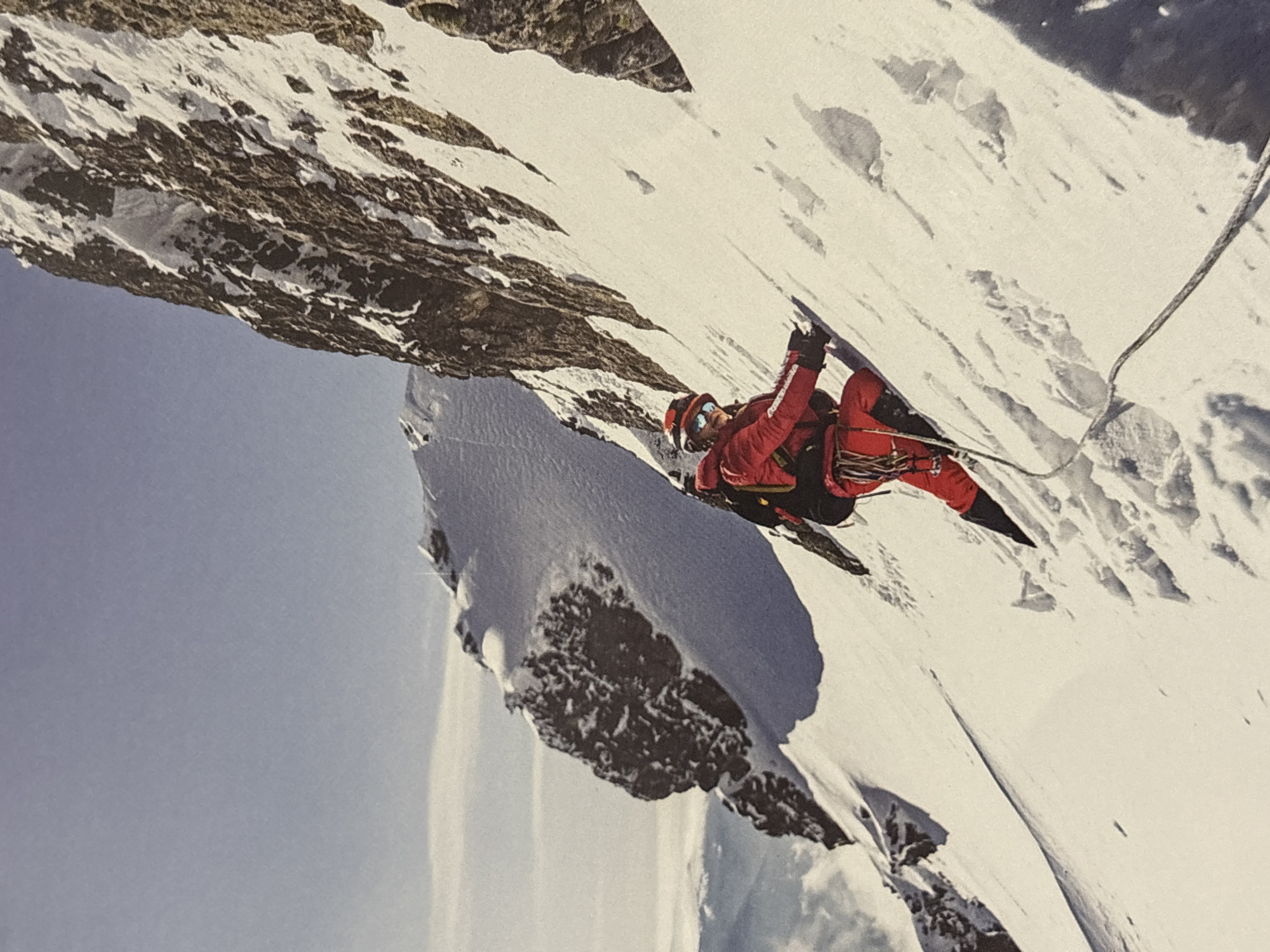 A lone mountain climber ascends a steep, snow-covered slope. The climber is clad in a red snowsuit, a backpack, goggles, and a helmet. They are using their hands and feet to grip the icy surface. A rope extends from the climber down the slope, suggesting they are part of a climbing expedition. The environment is a rugged mountain landscape with snow-capped peaks and a clear blue sky. The scene is lit by bright sunlight, casting sharp shadows on the snow. The image is a photograph, likely taken by another climber or from a vantage point on the mountain. There is no discernible connection to the city of Plovdiv, Bulgaria.