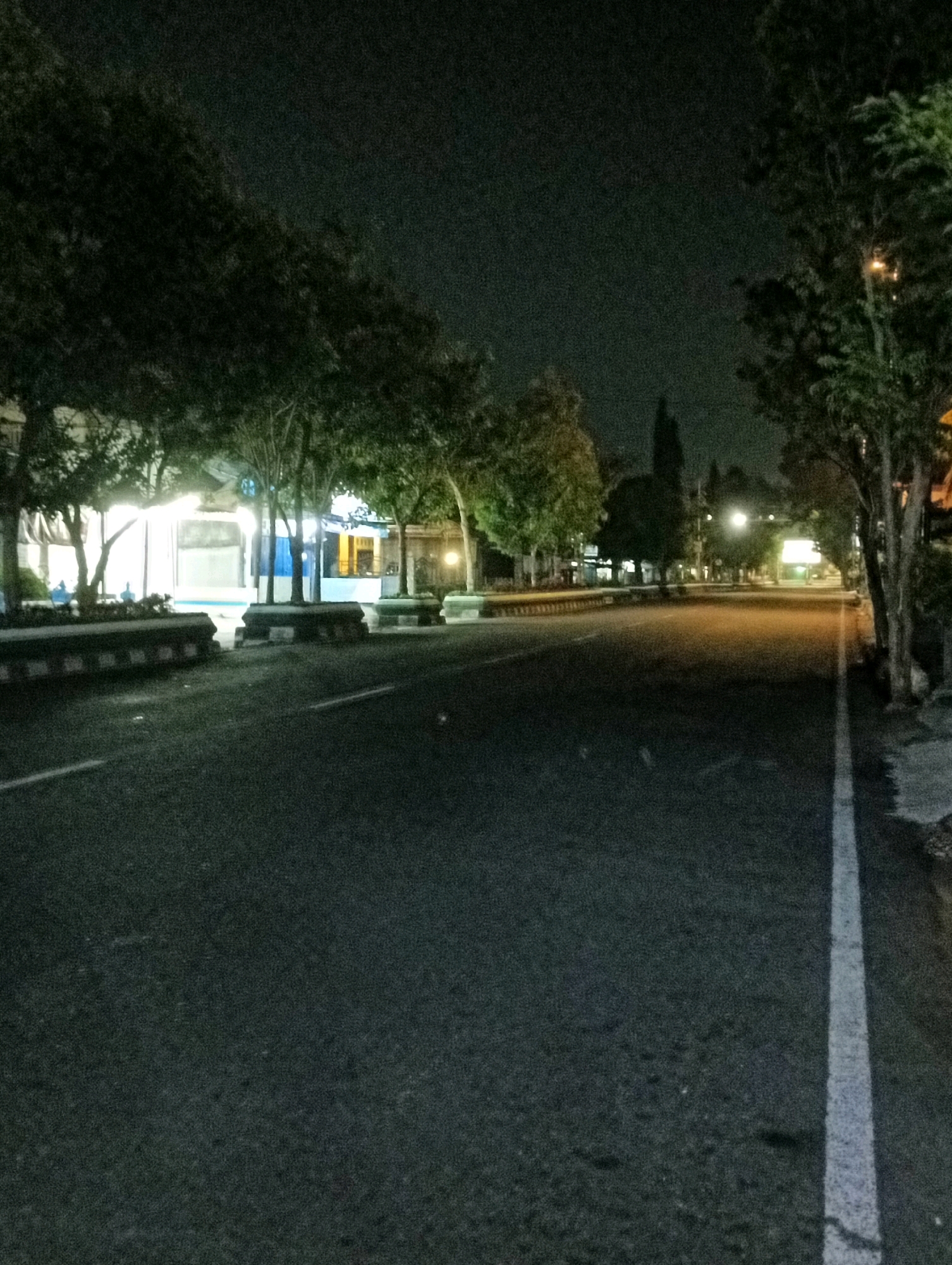 This image captures a street scene at night in Kliwonan, Indonesia. The asphalt road is dimly lit, with a white line marking the edge of the lane on the right. On the left side of the road, there are trees and buildings with interior lights illuminating the facade. Some concrete barriers with reflective markings are visible along the roadside. The sky is dark, suggesting it is late at night or early morning. No people or vehicles are immediately discernible in the frame. The overall impression is of a quiet, possibly urban or suburban, street after dark.