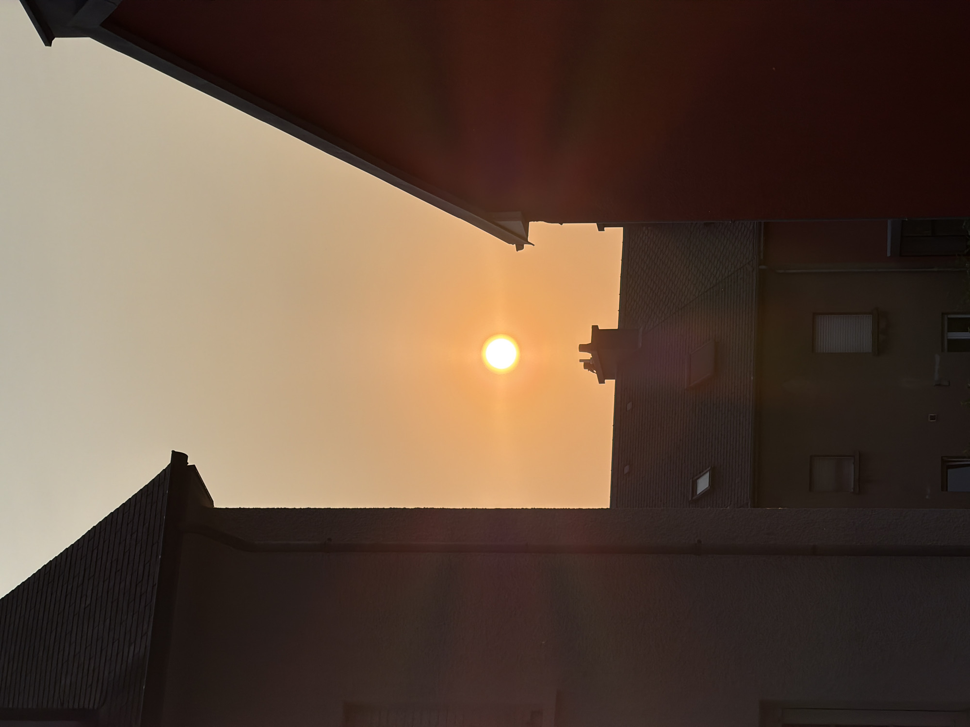 A bright, hazy sun hangs in the pale orange sky, positioned centrally in the frame, slightly above the rooftops of residential buildings. The sun is a distinct, glowing orb with a soft halo of light surrounding it. Two houses, one on the left and one on the right, partially frame the view of the sun and the sky. The house on the left is dark, with a dark, tiled roof sloping towards the right. The house on the right is a deep reddish-brown color, also with a dark roofline visible. In the middle ground, between the two foreground houses, are more buildings with dark tiled roofs. One of these buildings has a visible chimney. The overall scene is one of a hazy sunset or sunrise over a built-up area. The image does not contain any people, animals, or specific actions taking place. The location in Esch-sur-Alzette, Luxembourg, is a residential urban environment.