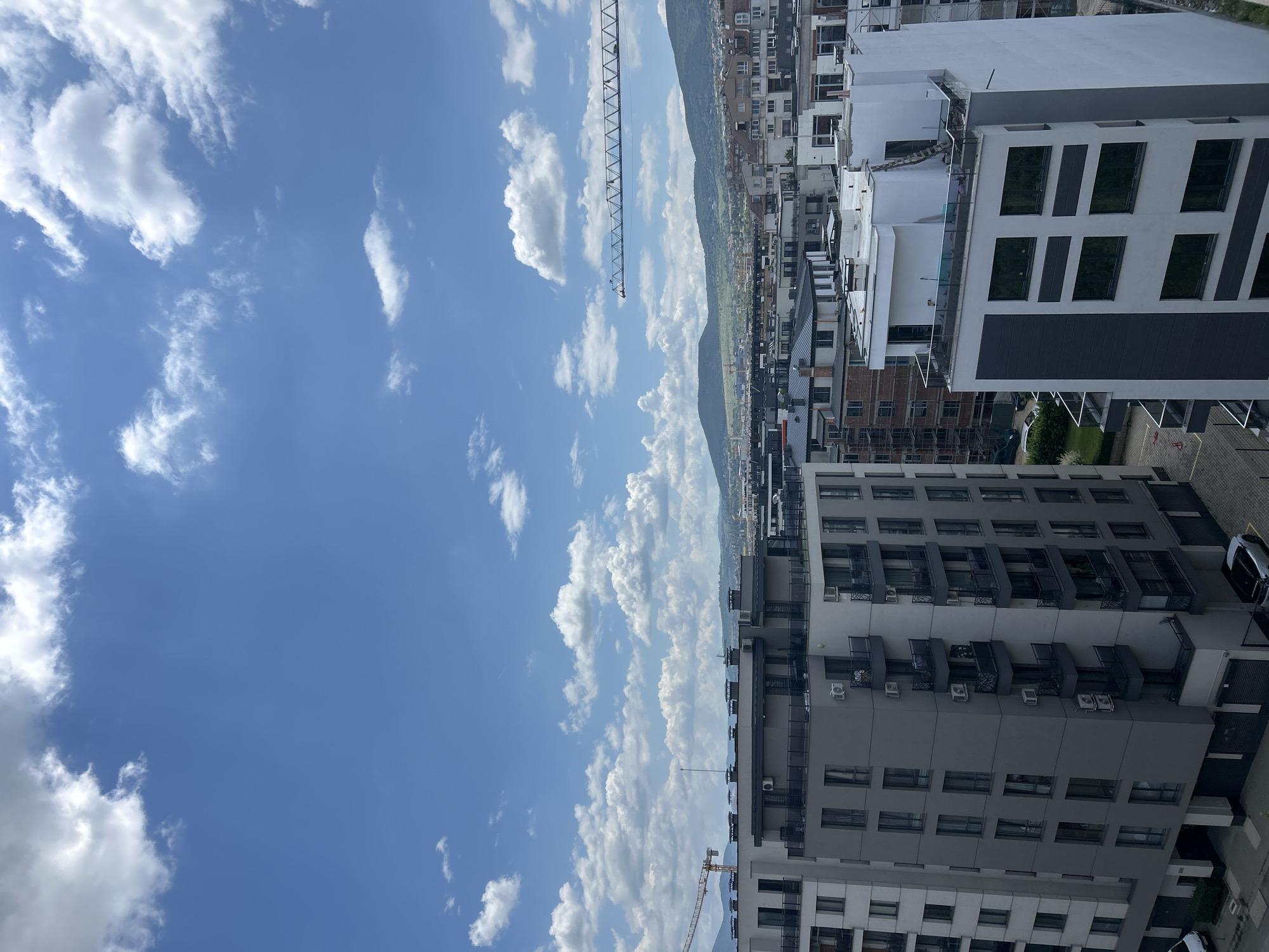 This is an outdoor, eye-level, vertical shot of a city skyline in Sofia, Bulgaria, under a bright blue sky with scattered white clouds. In the foreground, a modern, multi-story apartment building dominates the left side of the frame. It features a grey facade with dark-framed windows and balconies, and several air conditioning units visible on the exterior. To the right of this building is another apartment building, predominantly white with dark architectural accents and large windows. The background reveals a dense urban landscape with various buildings of different architectural styles, some appearing to be under construction, indicated by scaffolding and cranes. Further in the distance, rolling green hills form a natural backdrop to the city. The overall impression is one of urban development and contemporary architecture against a scenic natural setting.
