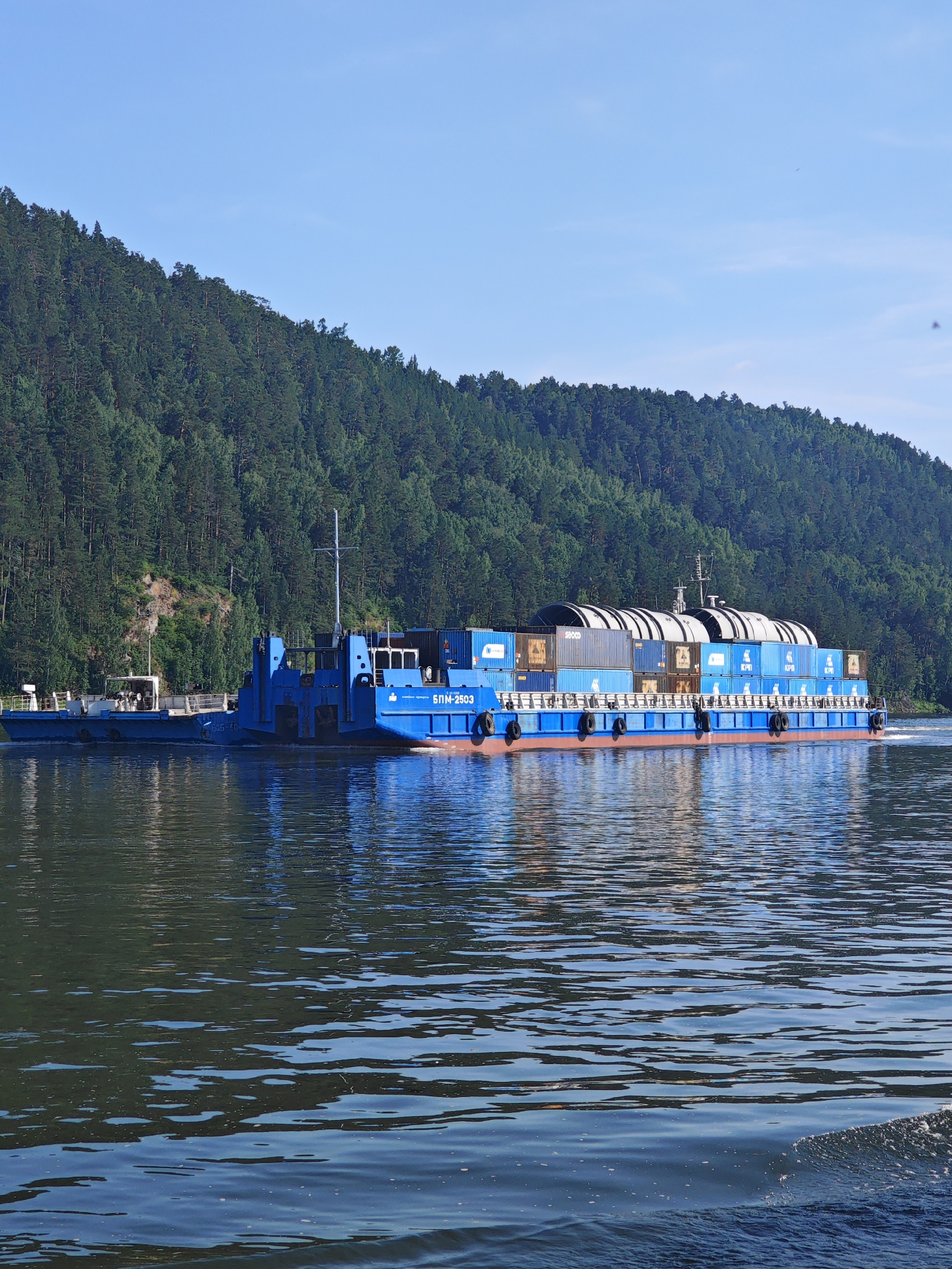 The image shows a large blue barge carrying numerous shipping containers and large cylindrical objects on a wide river. The barge is moving from left to right. In the background, a dense green forest covers a steep hillside, and a clear blue sky is visible above. The water in the foreground is dark and reflects the trees and sky, with some small waves and foam visible on the right side. A smaller, lighter-colored barge is attached to the left of the main barge. The main barge has Cyrillic lettering on its side that reads "БЛМ-2503". The scene appears to be of the Yenisei River near Predivinsk, Russia, a town known for its river port and transportation of goods along the waterway.
