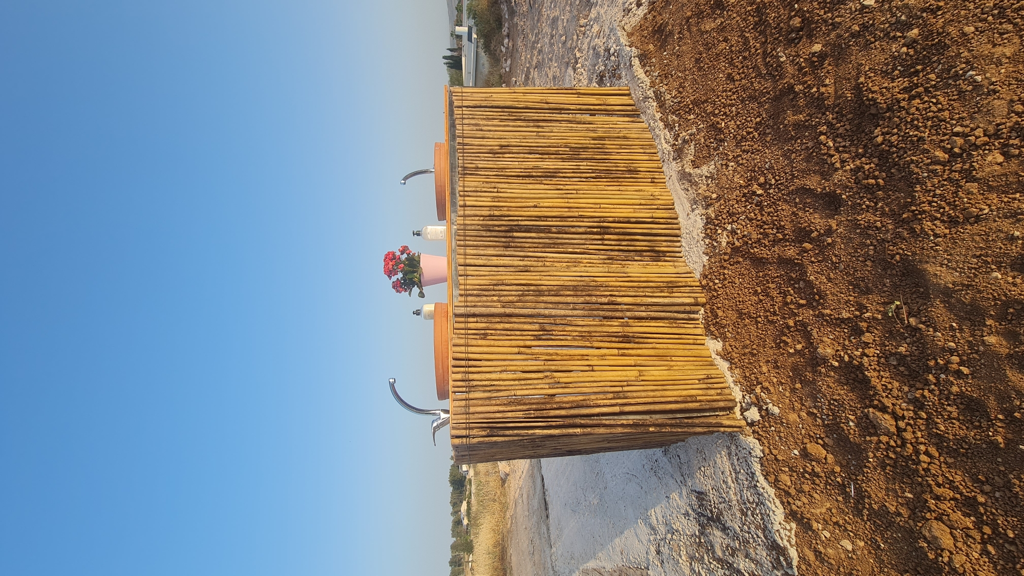 The image shows a rustic outdoor vanity made of bamboo, situated in the Lachish Regional Council, Israel. The vanity has two chrome faucets mounted on wooden countertops. A pink pot containing red flowers sits on one of the countertops. The vanity is placed on a cement or concrete base, adjacent to a patch of dry, rocky ground. The background includes a clear blue sky. In the distance, there is a glimpse of greenery and possibly some structures. The photograph appears to have been taken during daylight hours with good lighting.