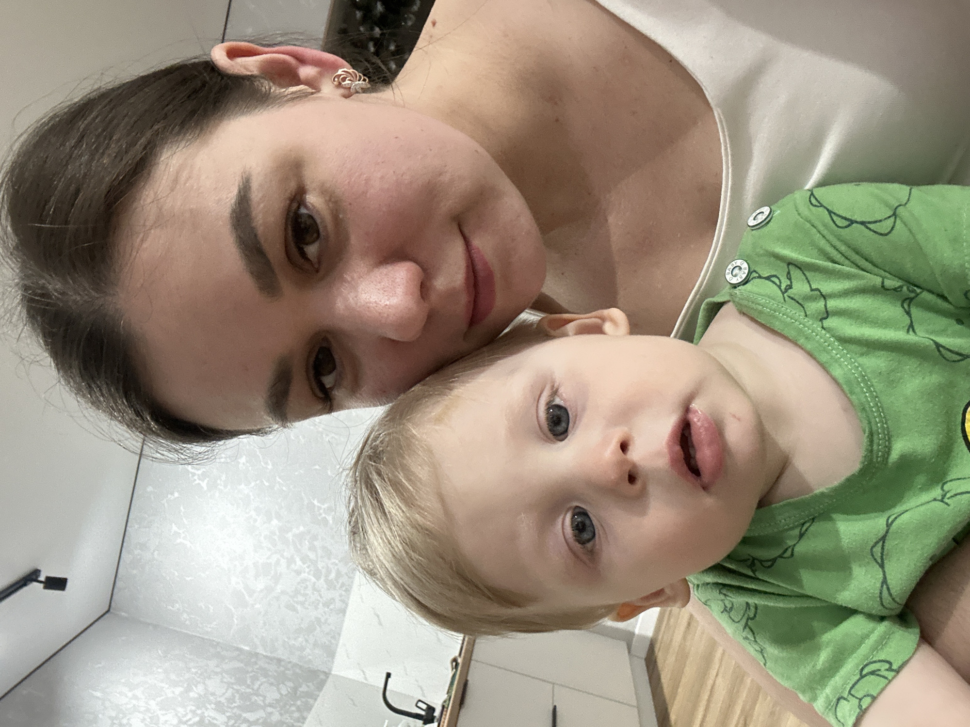 A woman with dark hair and fair skin is holding a young child with blonde hair and blue eyes. The woman is wearing a white top, and the child is wearing a green shirt. The background appears to be a room with white walls and wooden floors. Based on the available information, there is no indication this photo was taken in Chaikovsky, Russia.