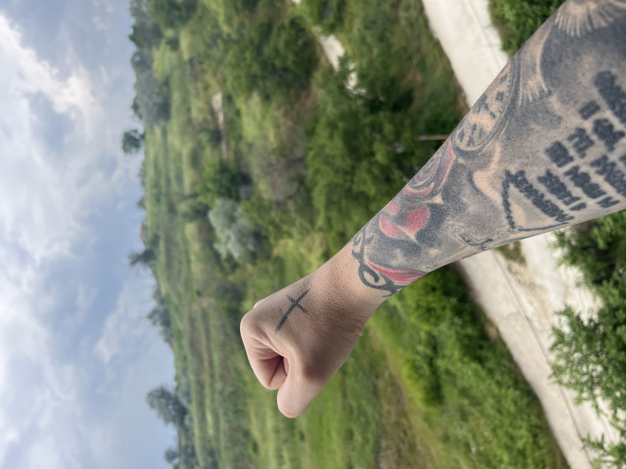 A forearm and hand are presented against a blurred background. The forearm is extensively tattooed. The hand is held in a fist with a cross tattoo. The background consists of a slope covered in trees and grass, and a path. It is not possible to ascertain the specific location in Varna, Bulgaria, without more details.