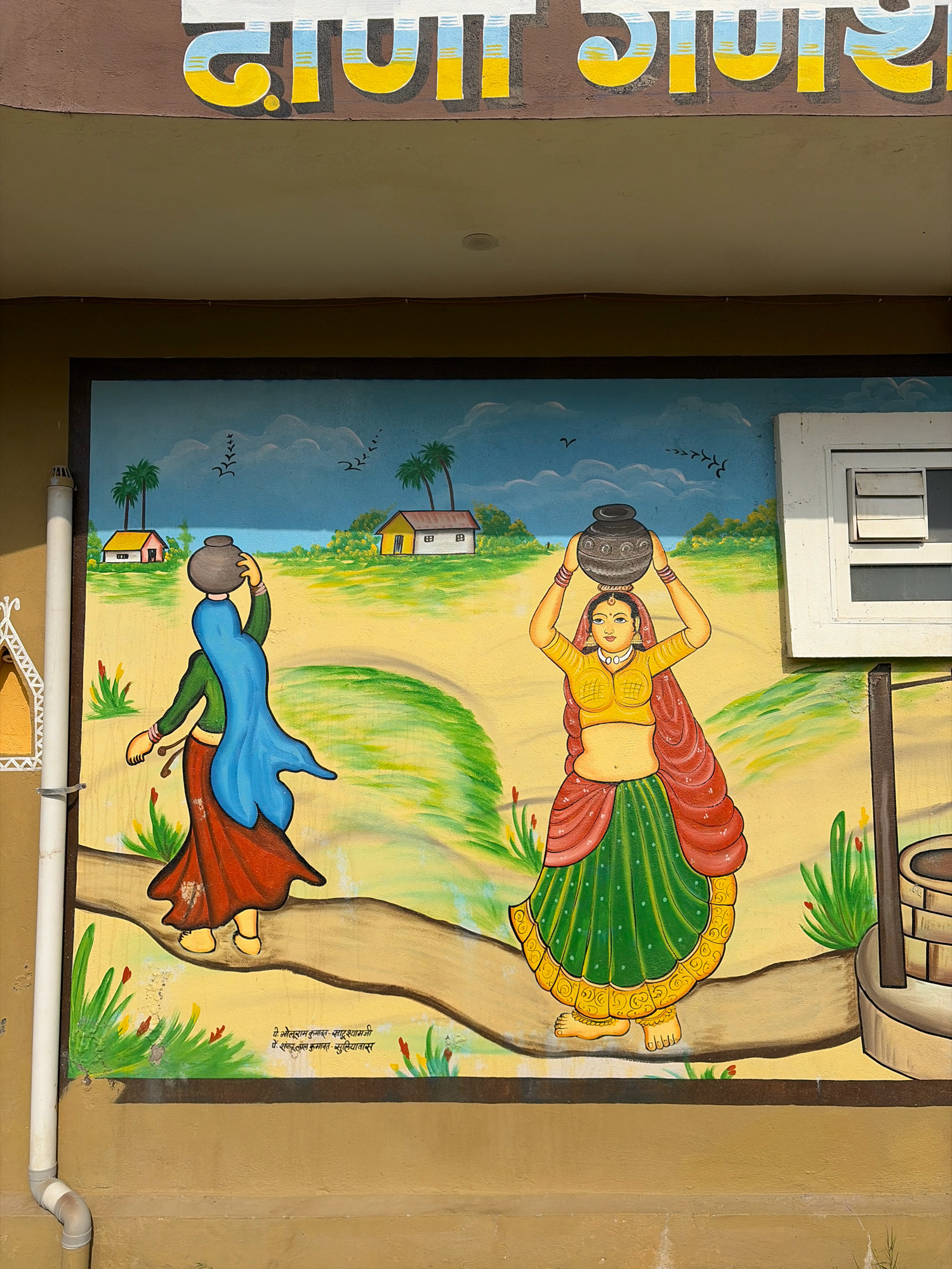 This image depicts a colorful mural painted on the exterior wall of a building in Dockiya, India. The mural features a rural Indian landscape with two women carrying water pots on their heads. The woman on the left wears a green blouse and a red skirt, with a blue veil covering her head and shoulders. The woman on the right is adorned in a yellow blouse, a green skirt, and a red shawl. Both women are walking along a dirt path. The background includes two houses with yellow roofs, palm trees, and a blue sky with birds. The mural is bordered by a dark frame. Text in Hindi is visible at the top of the image and at the bottom of the mural.