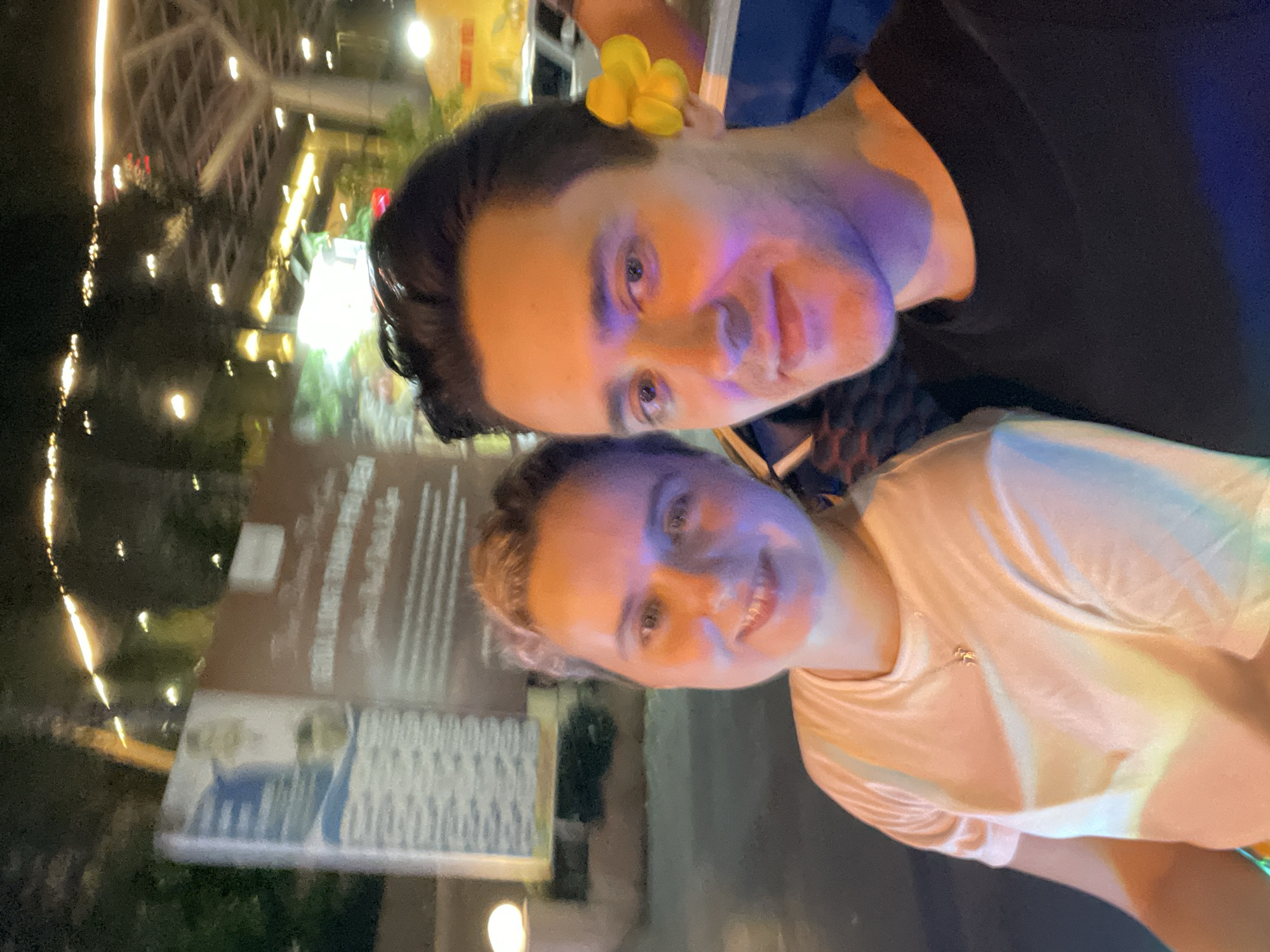 This image features two individuals, a male and a female, posing together in a nighttime outdoor setting in Nha Trang, Vietnam. The male is in the foreground, facing the camera with a gentle expression. He has dark hair and is wearing a black shirt. A bright yellow plumeria flower is charmingly tucked behind his right ear. His face is distinctly illuminated by multi-colored lights, casting blue and purple hues on one side and warmer orange tones on the other.
Behind him and slightly to his right is a smiling female, also looking towards the camera. She has lighter hair and wears a light-colored top, possibly white or a very pale peach, with a small, delicate necklace visible around her neck. Her face and clothing are also highlighted by the ambient artificial lighting, creating various colorful reflections.
The background is blurred but clearly shows numerous bright artificial lights, indicating an active urban or tourist area at night. These lights suggest illuminated buildings, signs, and possibly streetlamps, contributing to a lively and vibrant atmosphere. While there are visible signs in the background, the text on them is indecipherable due to the blur. The overall mood is joyful and relaxed, conveyed by the happy expressions of both individuals.