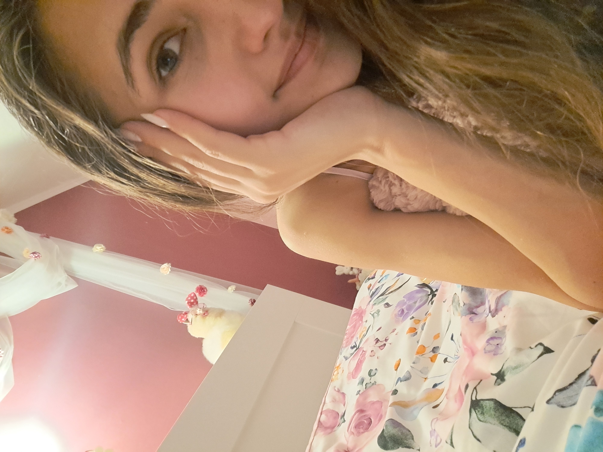 An overhead medium shot captures a young woman with long brown hair, resting her head on her hand as she looks toward the camera. Her face is framed by loose strands of hair, and she is wearing a light pink camisole. Behind her, a bed is covered with a floral duvet. In the background, a white canopy hangs from the ceiling, adorned with small, colorful decorations. A stuffed animal with pink polka-dot ears and glasses sits on the edge of a white shelf in the background. The room is painted a soft pink hue.