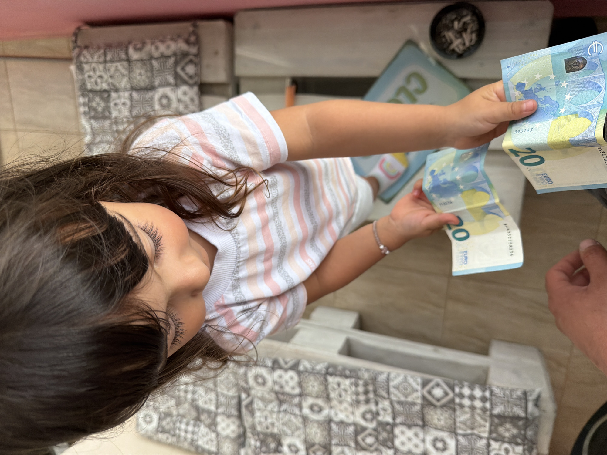 A high-angle shot shows a young girl, viewed from above, holding two 20 Euro banknotes in her hands. She is wearing a striped t-shirt with pink, white, and gray stripes and a bracelet on her left wrist. The girl is looking down at the money. In the background, there is a patterned bench, a pink wall, and a glimpse of a floor. There is also a small bowl containing what appear to be cigarette butts or ashes to the right of the girl. The scene appears to be indoors, possibly a residential setting. The image does not provide any specific visual cues to indicate a location in Burgas, Bulgaria.
