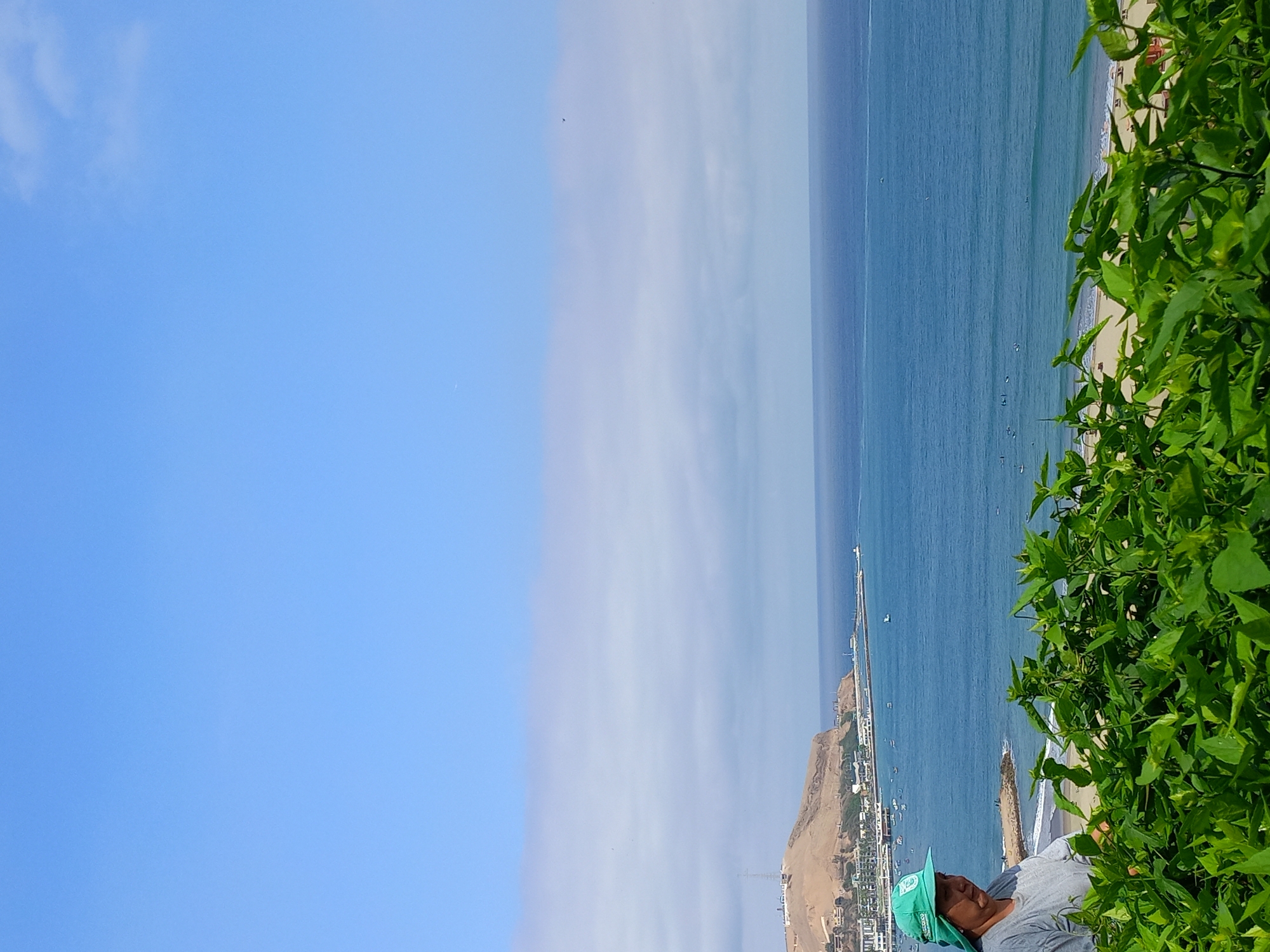 A woman wearing a green hat and a grey shirt stands on a hill overlooking the ocean in Barranco, Peru. She is partially obscured by lush green foliage in the foreground. In the middle ground, a beach is visible with several people in the water, some appearing to be surfing. Beyond the beach, a coastal city stretches along the shoreline, with buildings and a prominent hill topped with a structure that resembles a transmission tower. The ocean is a deep blue, and the sky is a clear, light blue with a few wispy clouds.