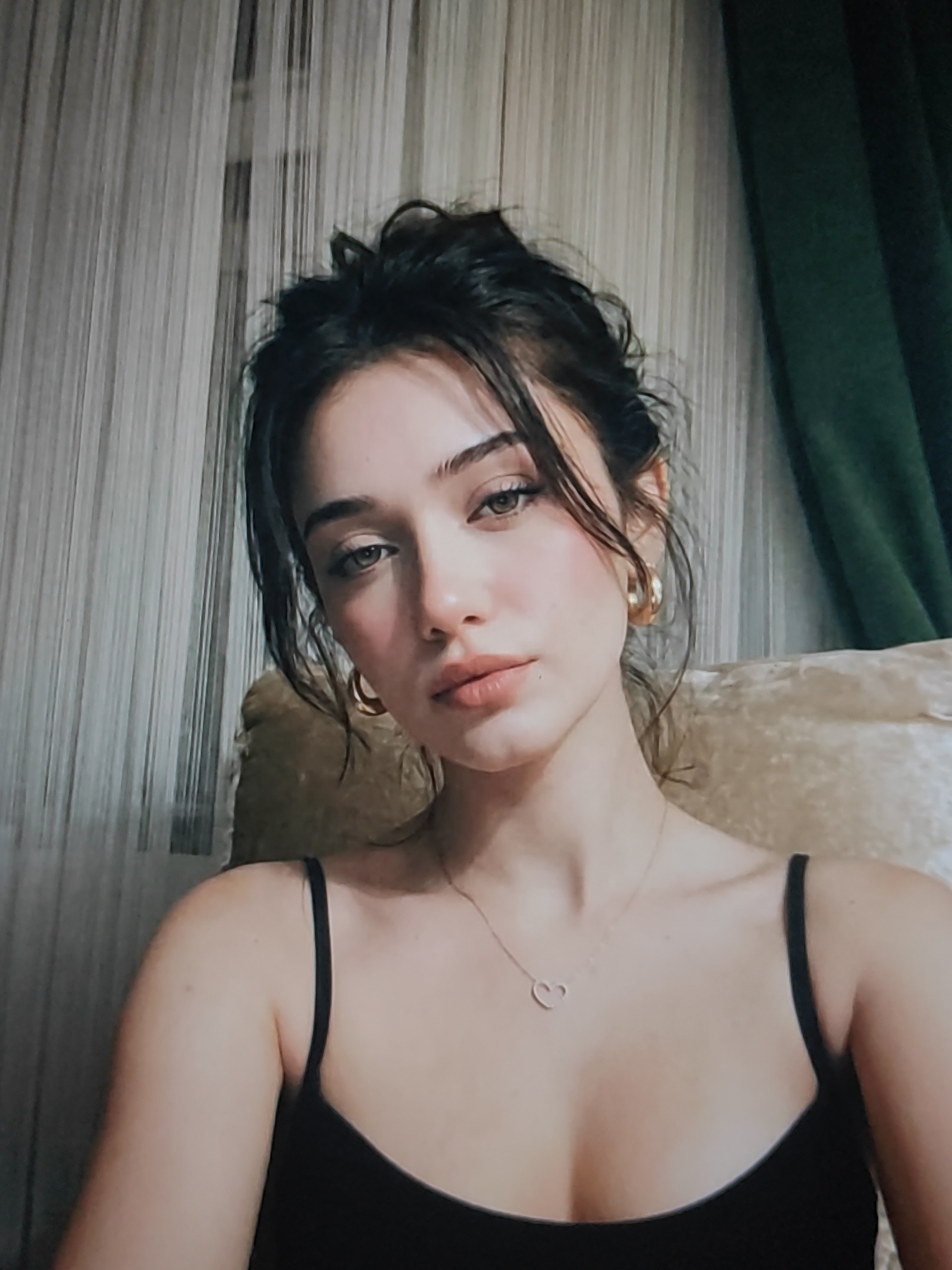 A close-up portrait of a young woman with dark, messy hair and light brown eyes is shown. She is wearing a black tank top and a delicate heart-shaped necklace. Her expression is soft and alluring, with a slight gaze towards the camera. Large hoop earrings adorn her ears. The background features vertical blinds and a hint of a green curtain, suggesting an indoor setting, possibly a home. The lighting appears to be soft, possibly from natural light, casting a warm glow on her skin. There are no visible text elements in the image.