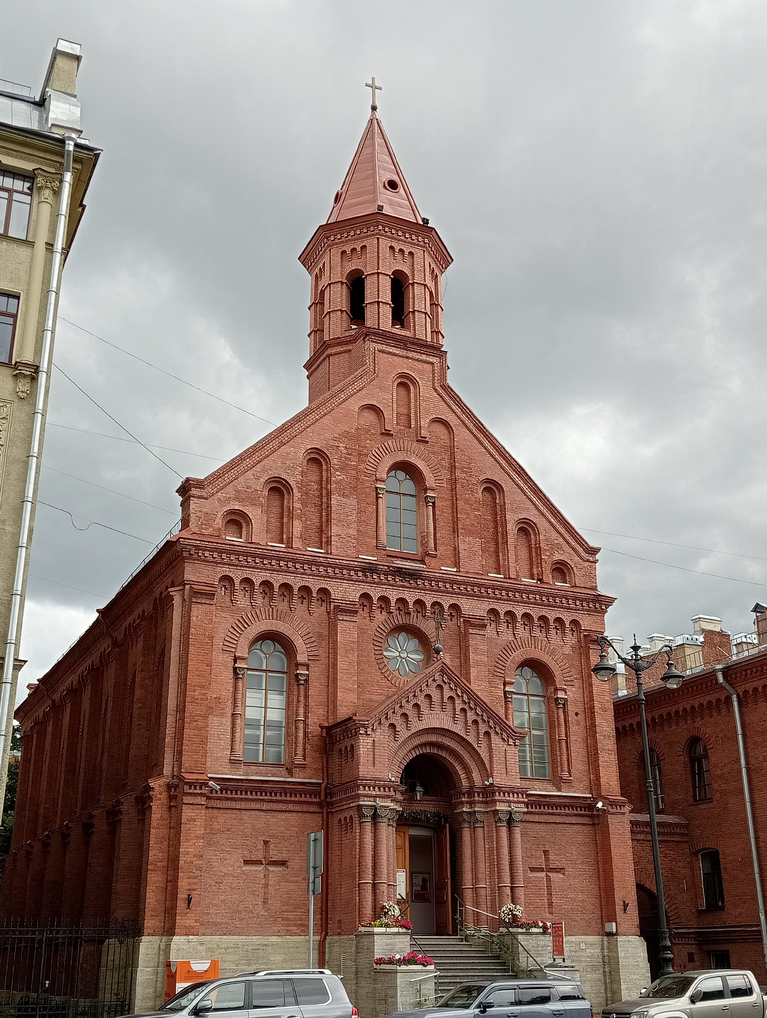 This is a ground-level, eye-level, medium shot of the Catholic Church of Saint Stanislaus in Saint Petersburg, Russia, on a cloudy day. The church is constructed of red brick and features a prominent bell tower topped with a cross. The facade has arched windows, decorative brickwork, and a central entrance with a portico. Steps lead up to the entrance, with flower arrangements on either side. Two cars are parked in front of the church on a paved surface, and a black metal fence runs along the property line. To the left of the church is a lighter-colored building with a metal downspout. Streetlights and overhead wires are also visible.