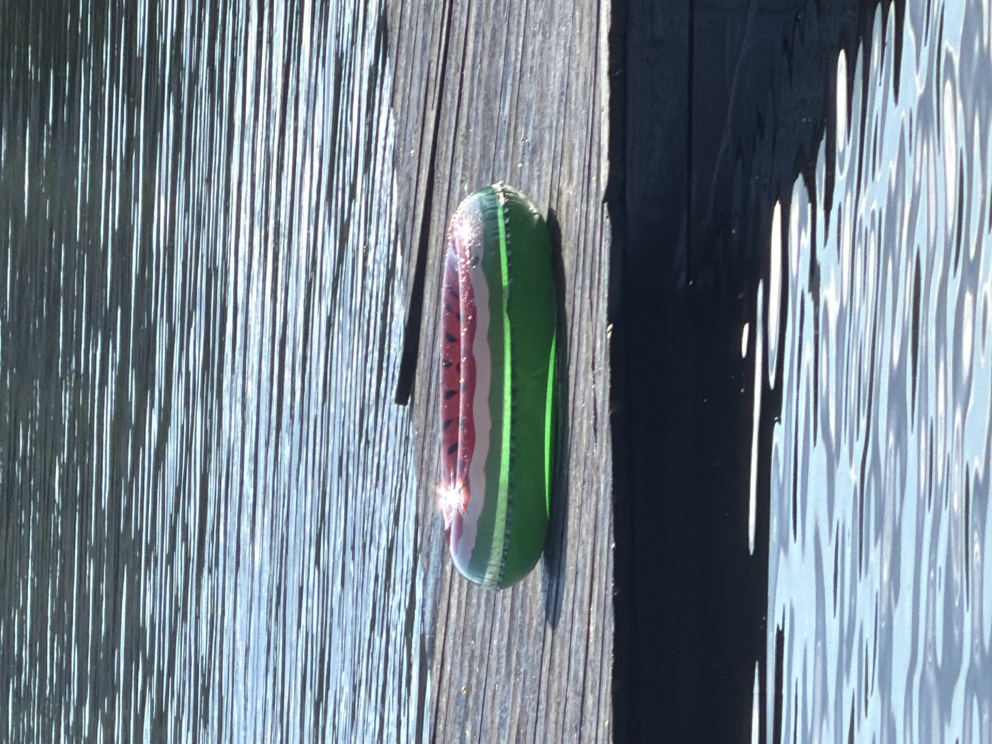 The image shows a glass pickle ornament resting on a wooden surface. The ornament is elongated and has vertical stripes in shades of green and purple, with a reflective surface that catches the light. The wooden surface appears to be a dock or pier, with visible grain and texture. To the left and right of the wooden surface are reflections of water, indicating the ornament is near a body of water. The lighting suggests it is daytime, with bright sunlight creating highlights on the ornament and the water. There is no text visible in the image.