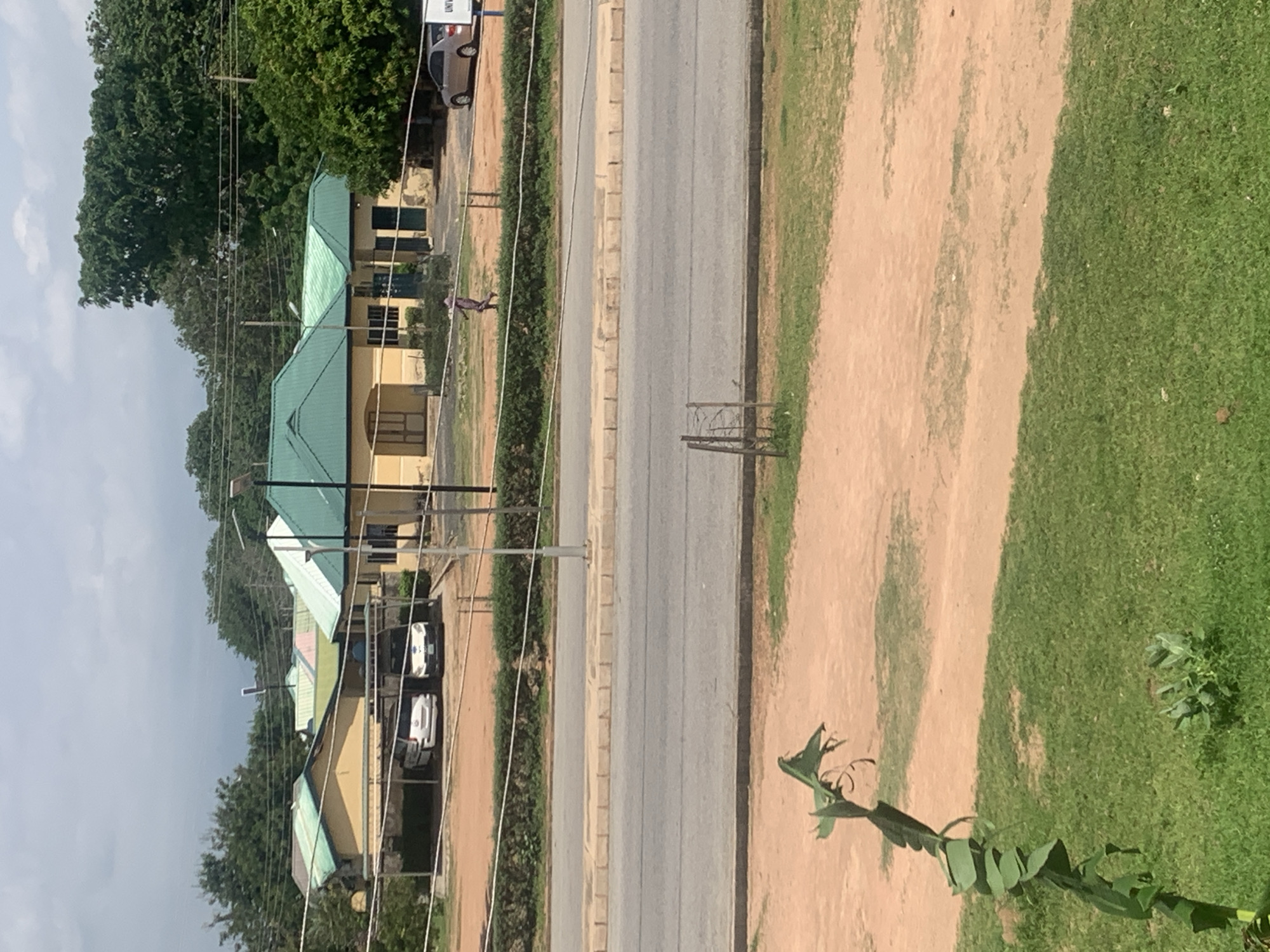 This image depicts an outdoor scene in Jalingo, Nigeria, featuring a road in the foreground and a cluster of buildings with green roofs in the background. A person in a purple garment is walking on a paved area between the road and the buildings. Several vehicles are parked under covered structures near the buildings. The environment includes lush green trees, a dusty patch of ground, and a stretch of asphalt road. The sky is overcast with scattered clouds.