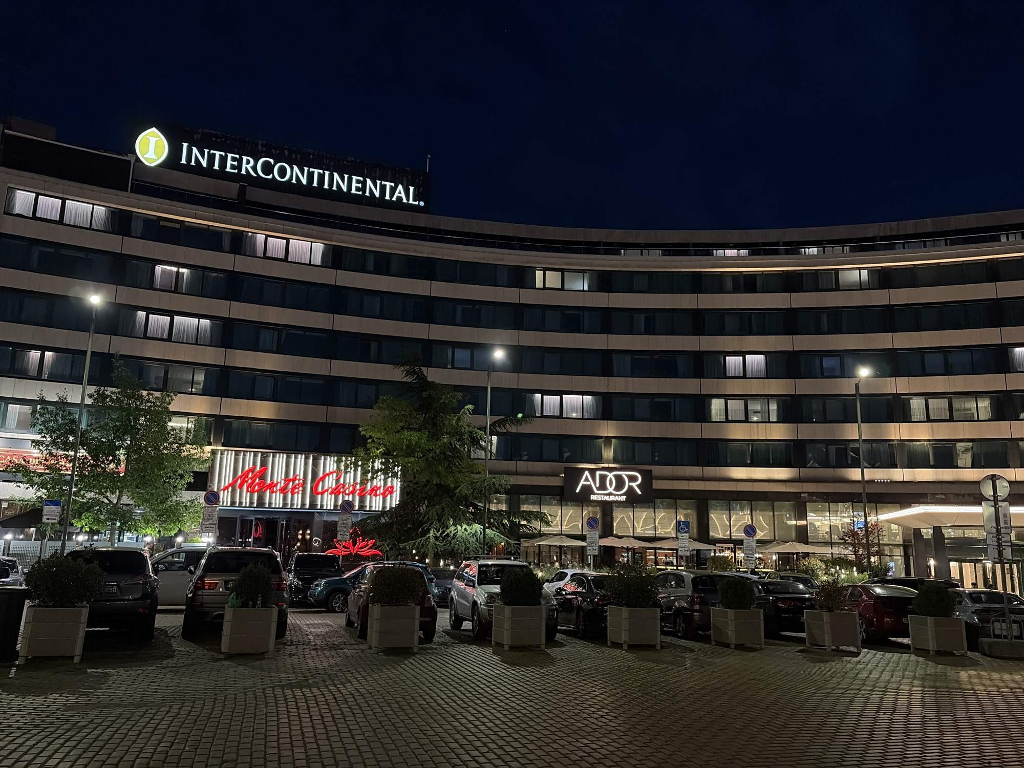 The image captures a nighttime exterior view of the InterContinental Hotel in Sofia, Bulgaria. The central subject is a large, multi-story building with a distinctive concave, curved facade. The hotel's illuminated "INTERCONTINENTAL" sign, featuring its "I" logo, is prominently displayed on the upper part of the structure. Numerous rectangular windows line the building's floors; some are lit from within, revealing white curtains, while others are dark.
At ground level, two establishments are visible: "Monte Casino" on the left, identified by a bright red neon sign and a wide glass entrance; and "ADOR RESTAURANT" to the right, marked by a white illuminated sign and a glass-fronted dining area with outdoor seating shaded by umbrellas.
The immediate foreground is a paved surface, textured with a block or cobblestone pattern, serving as a driveway or parking area. A selection of vehicles, including various SUVs and sedans of different colors, are parked or positioned along this area. Several light-colored, rectangular planters containing green shrubs are strategically placed. Multiple streetlights provide artificial illumination for the foreground and parts of the building. Green trees are interspersed, particularly near the casino and restaurant entrances. The sky above is dark blue, indicative of the nighttime setting. While no specific individuals are distinctly visible, the illuminated businesses and parked cars suggest ongoing activity within the premises. The scene depicts an upscale urban environment.