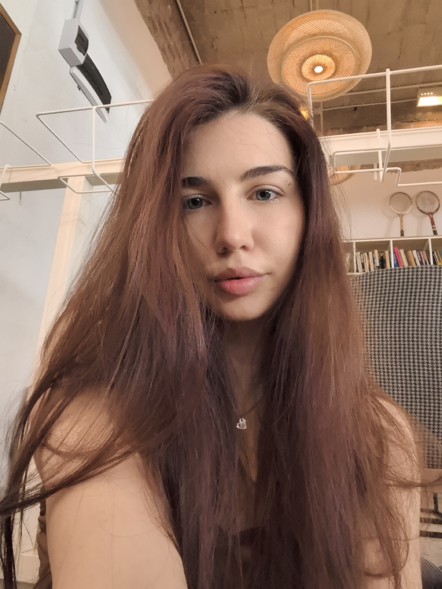 A close-up, eye-level shot features a woman with long, auburn hair and fair skin. She has blue eyes and light pink lips, and a delicate silver necklace is visible around her neck. The background reveals an interior space with a blend of modern and rustic elements. There is a white, metal framework of shelves, and a woven lampshade hangs from the ceiling. A bookshelf filled with books and two badminton rackets are visible. The location is potentially a cafe or a similar establishment in Stara Zagora, Bulgaria.