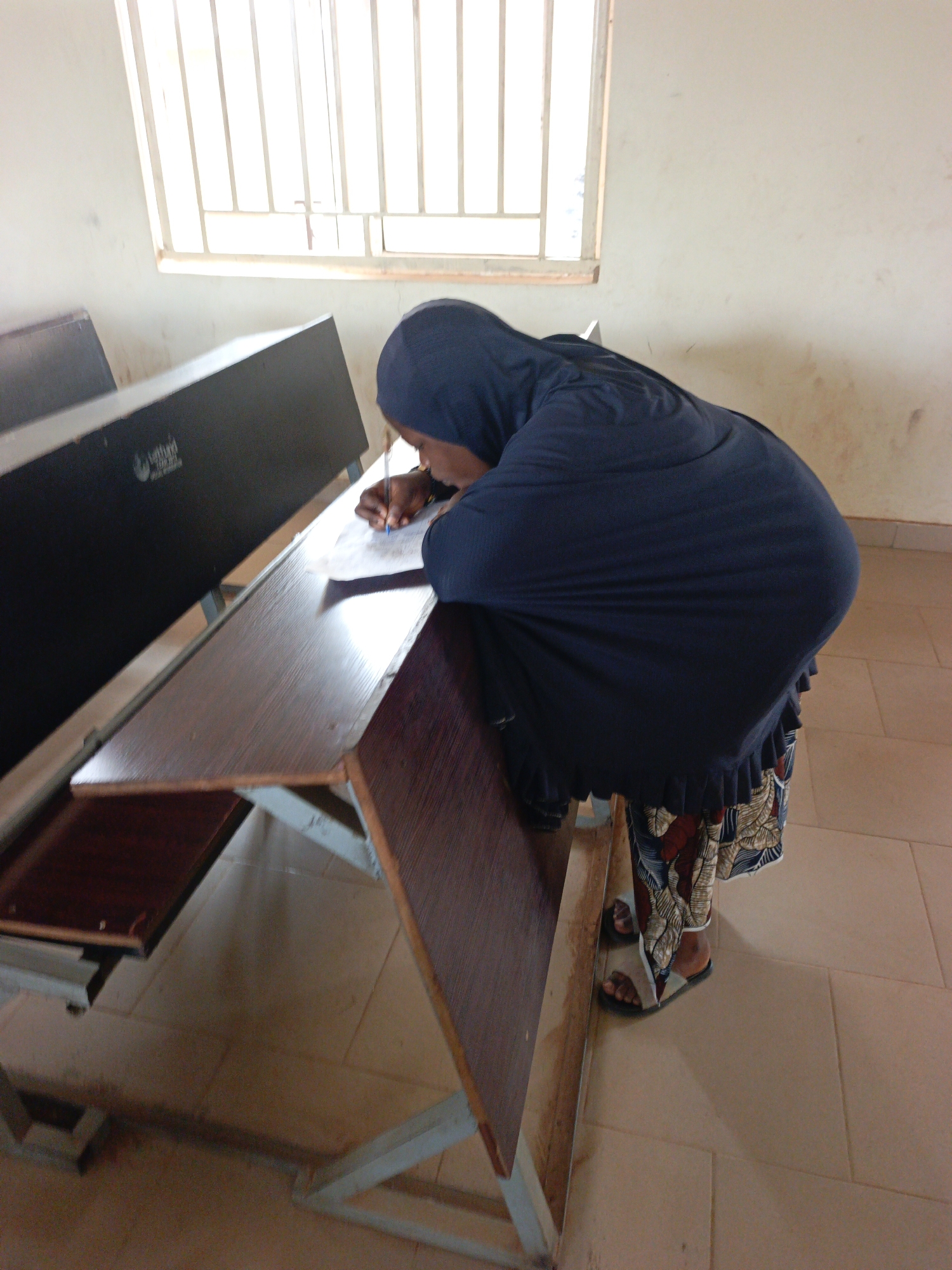 An individual wearing a dark blue hijab and a patterned skirt is leaning over a desk to write on a piece of paper. The desk is attached to a row of desks with metal legs in what appears to be a classroom or lecture hall. A window with a barred frame is visible in the background. The location is inferred to be in Akwanga, Nigeria.