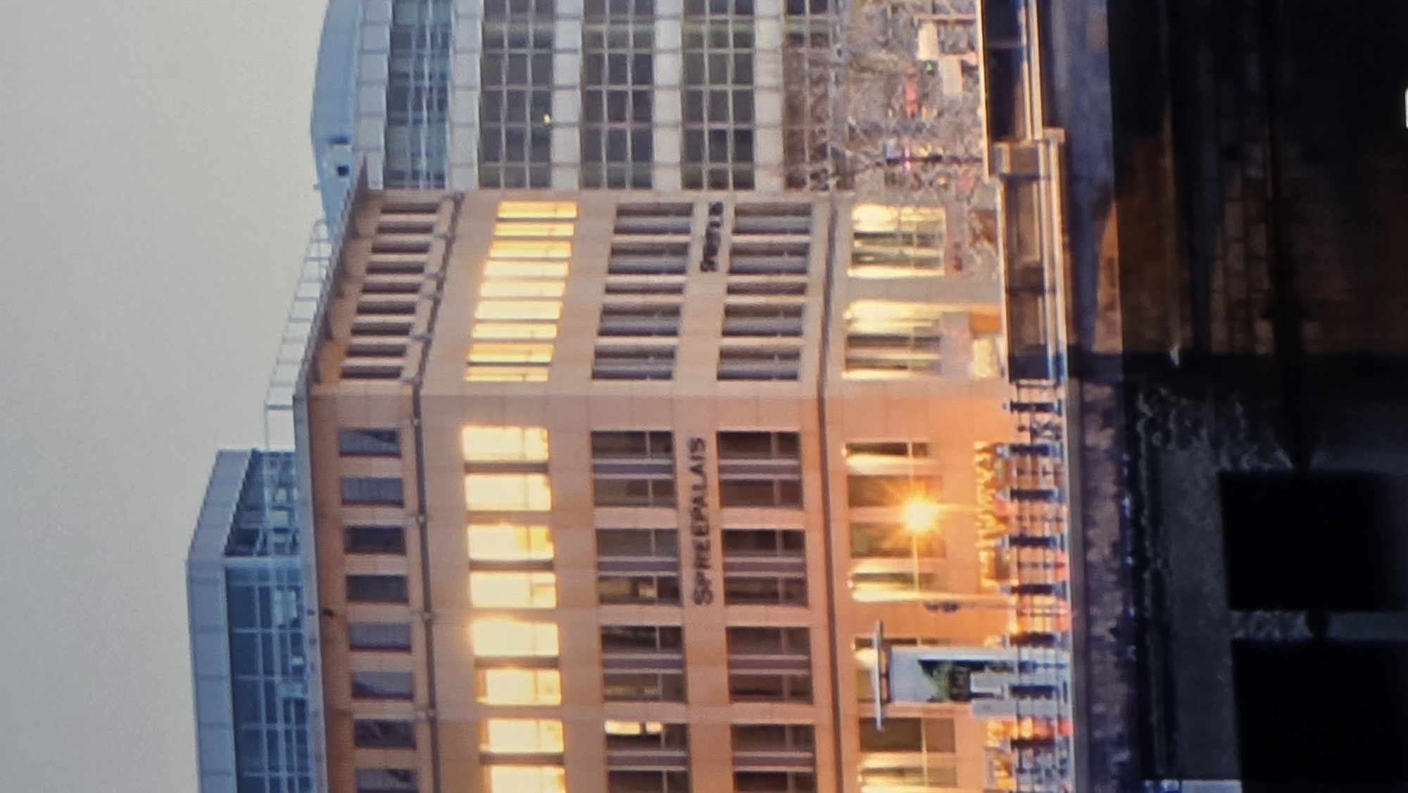 The image presents an urban waterside evening or early morning scene, located in the city of Dobrich, Bulgaria. A prominent, light-colored, multi-story building occupies the central frame. The building features numerous rectangular windows, with those on its upper floors reflecting a vibrant, golden light. On its facade, the name "SPREEPALAIS" is clearly inscribed in dark capital letters, and a partial inscription, possibly "SPREEPLAIS", is visible on a corner section.
To the left of the central building, a structure with a distinct glass-enclosed upper level is visible. To the right, a taller, modern building with a repeating grid pattern of windows extends into the background.
In the lower portion of the image, a dark body of water, likely a river or canal, is present. An embankment or bridge borders the water, equipped with a dark, decorative railing. A bright, circular streetlamp emits a strong, warm light near the water's edge, creating reflections. The sky overhead is a uniform, pale grey. No individual people are distinctly visible within the scene.