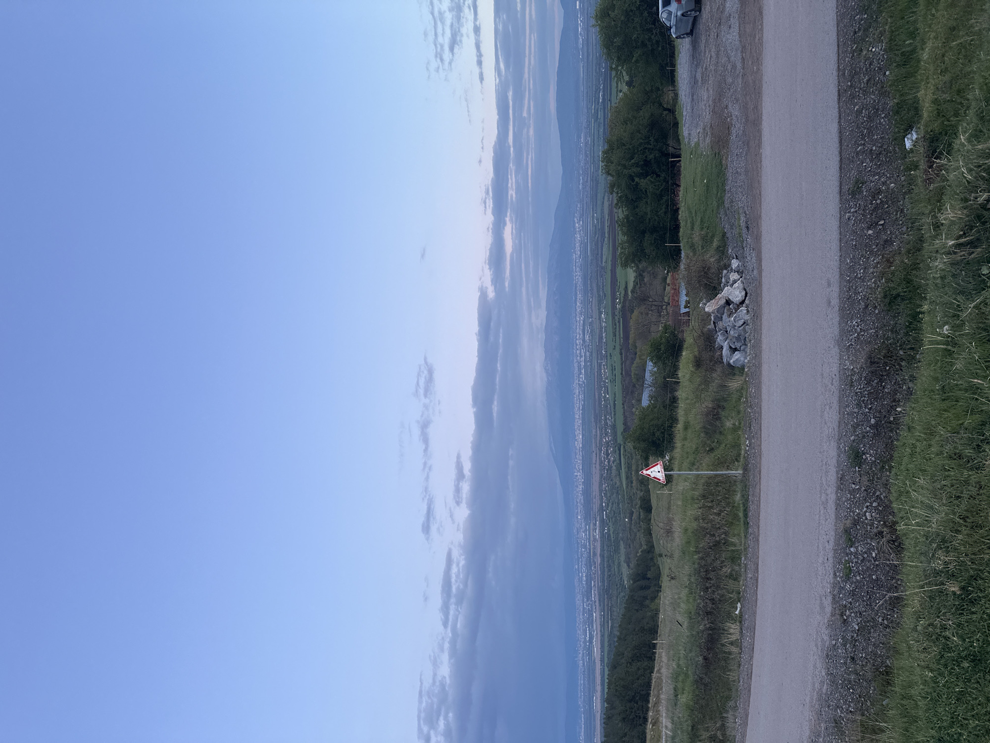 The image captures a panoramic landscape view of a valley and distant mountains in Balsha, Bulgaria, during twilight, suggesting either early morning or late evening. The sky, occupying the upper portion, is predominantly clear and blue, transitioning to a lighter hue towards the horizon, with scattered, elongated clouds.
In the middle ground, an expansive green valley is visible, dotted with what appear to be distant settlements or a small city, marked by faint patterns of buildings. Beyond the valley, a chain of mountains rises, appearing hazy and soft against the horizon.
The foreground on the right side of the image features a partially unpaved or gravel road running through a grassy area. To the left of the road, a triangular warning sign with a red border, white interior, and a black exclamation mark stands on a pole. The terrain immediately surrounding the road consists of green grass, gravel, and scattered rocks. On the far right, near the top, a portion of a white vehicle, possibly a van or camper, is visible, parked near some trees. No specific activity or event is taking place; it is a static landscape scene. No discernible text is visible other than the exclamation mark symbol on the warning sign.