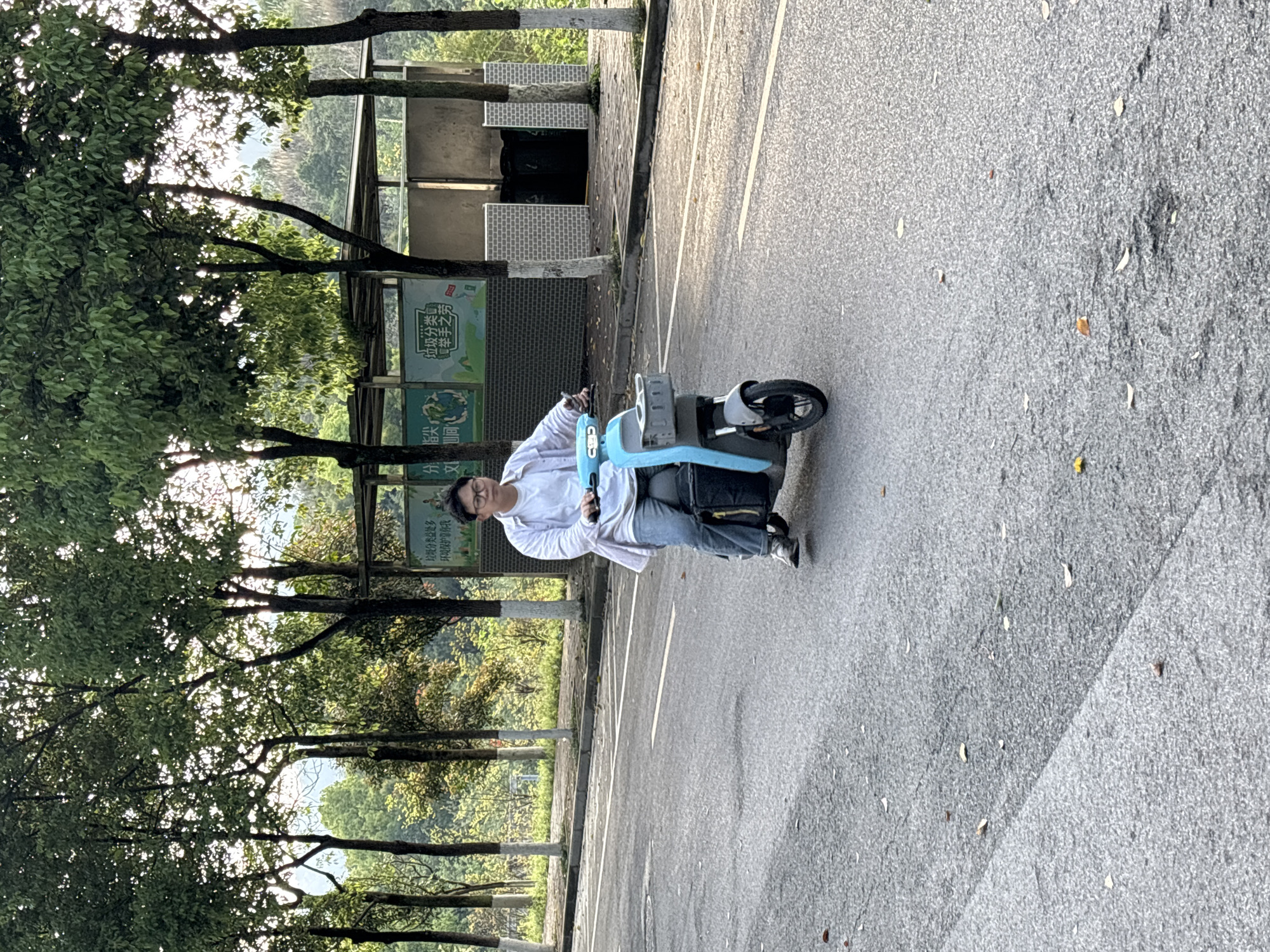 The image shows a person riding a light blue and gray electric scooter on an asphalt road. To the left, there is a park area with trees and a fence. A sign near the fence has Chinese characters on it, reading "垃圾分类益处多, 环境保护望你我" and "垃圾分类 举手之劳." Beyond the sign, there are black trash cans in a white enclosure. The background shows a glimpse of a distant hillside. The image was taken in the city of Jiangjin, China.