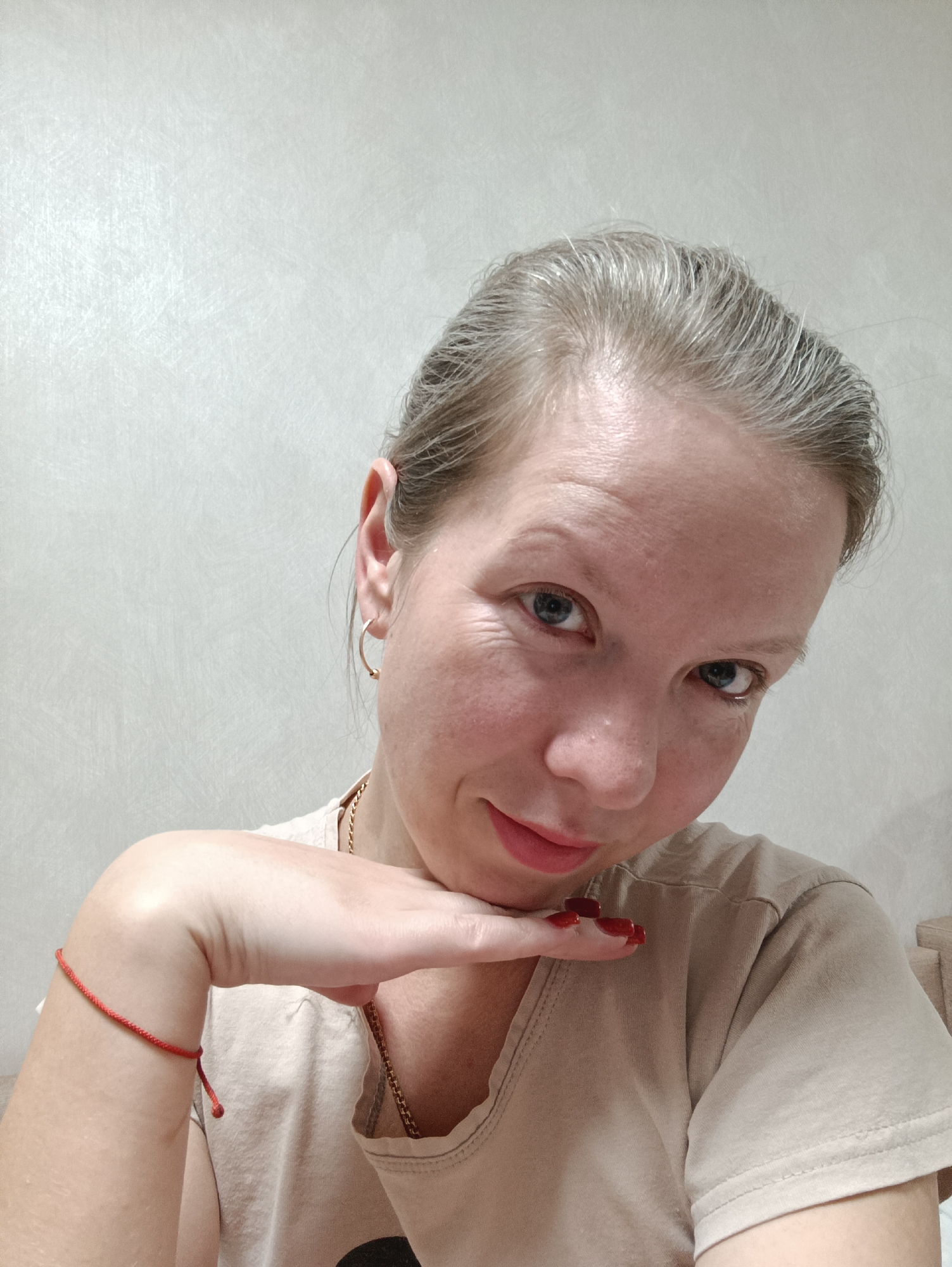 A woman is posing for a selfie. She has fair skin, blue eyes, and her graying hair is pulled back. She's wearing a light brown t-shirt and gold hoop earrings. A thin gold chain is visible around her neck. Her nails are painted a dark red. She's resting her chin on her hand, which has a red string bracelet. The background is a textured, light gray wall. There is no way to determine a specific location in Chaikovsky, Russia, from this image alone.
