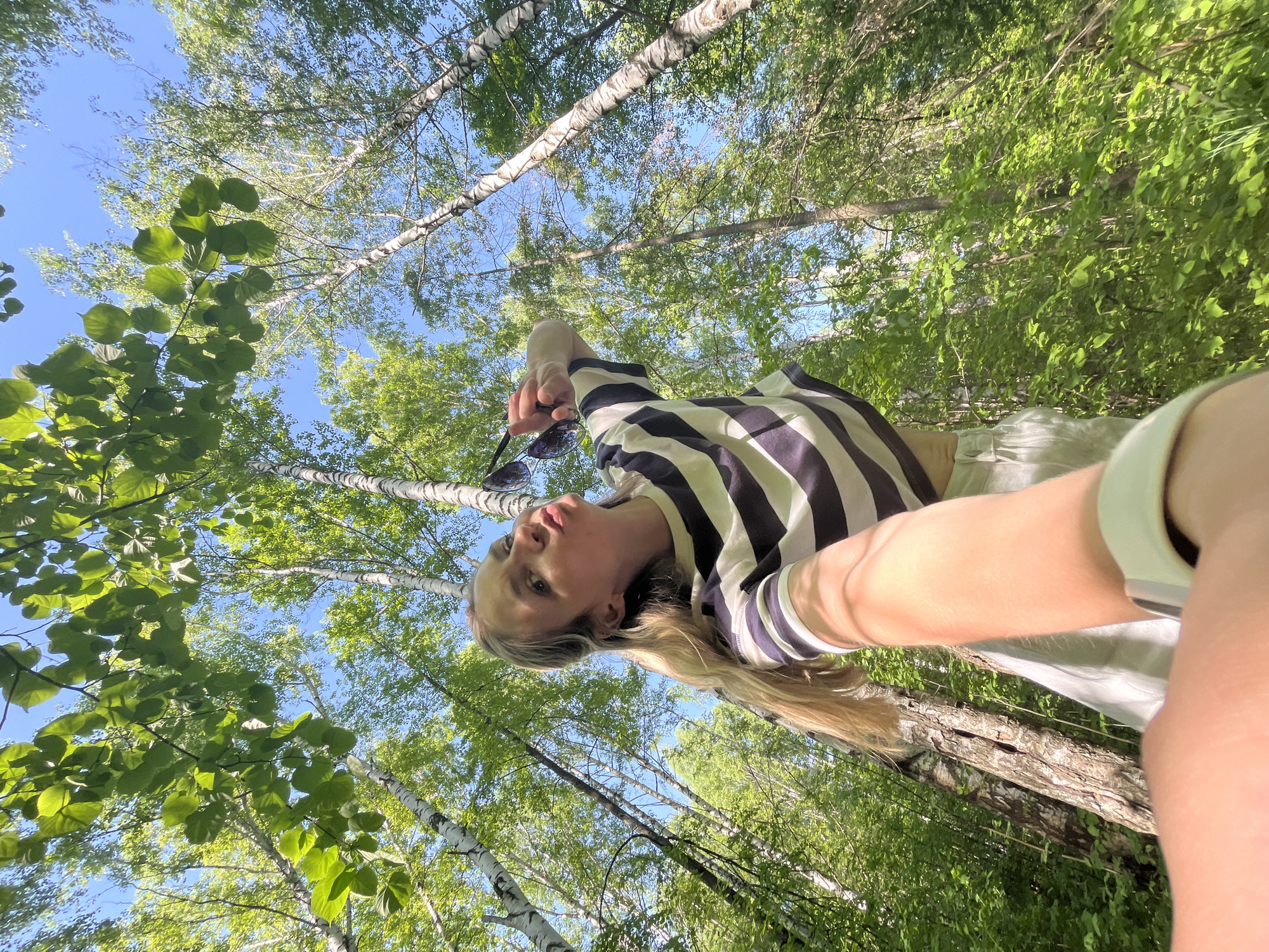 A low-angle selfie of a young woman with long blonde hair, holding sunglasses, is set against a backdrop of birch trees and green foliage under a blue sky. The photo appears to have been taken in a forest setting. Given the prompt, the specific location is inferred to be within the city of Chaikovsky, Russia, though visual confirmation is not possible. The woman is wearing a striped shirt. The trees are tall and the leaves are lush, indicating a possibly dense forest environment.